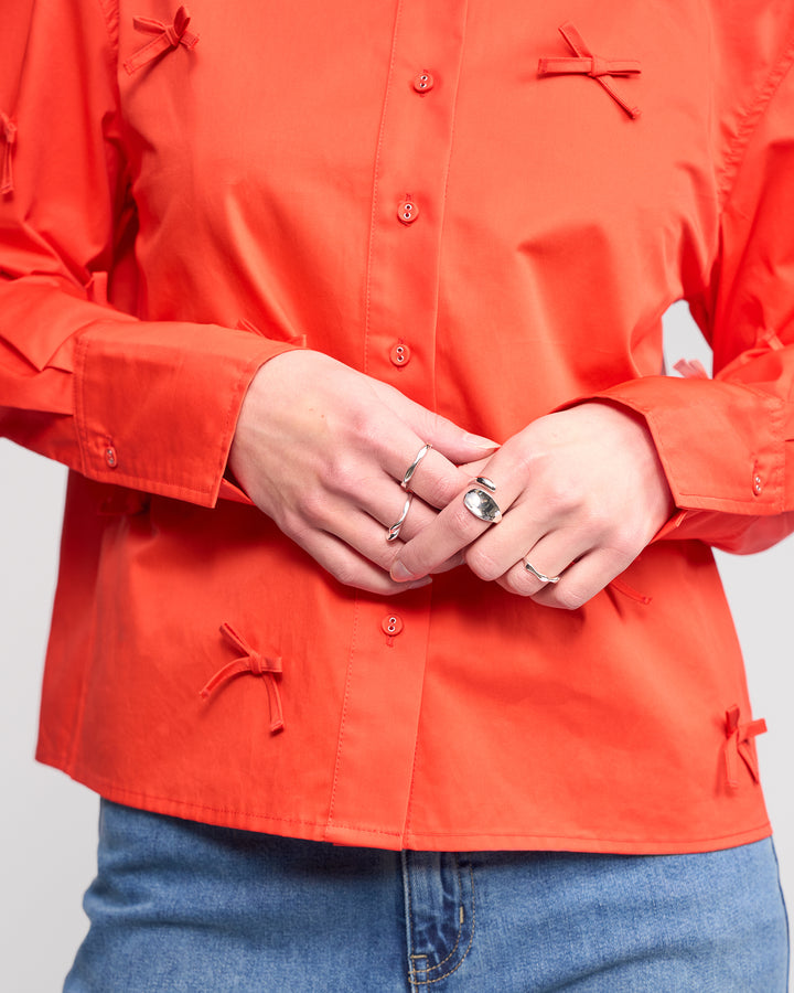 A person wears the Knewe Label Bow Shirt - Lava, a bright orange top with bow appliqués, paired with blue jeans. Their hands, adorned with several silver rings, are visible while their face is not shown.