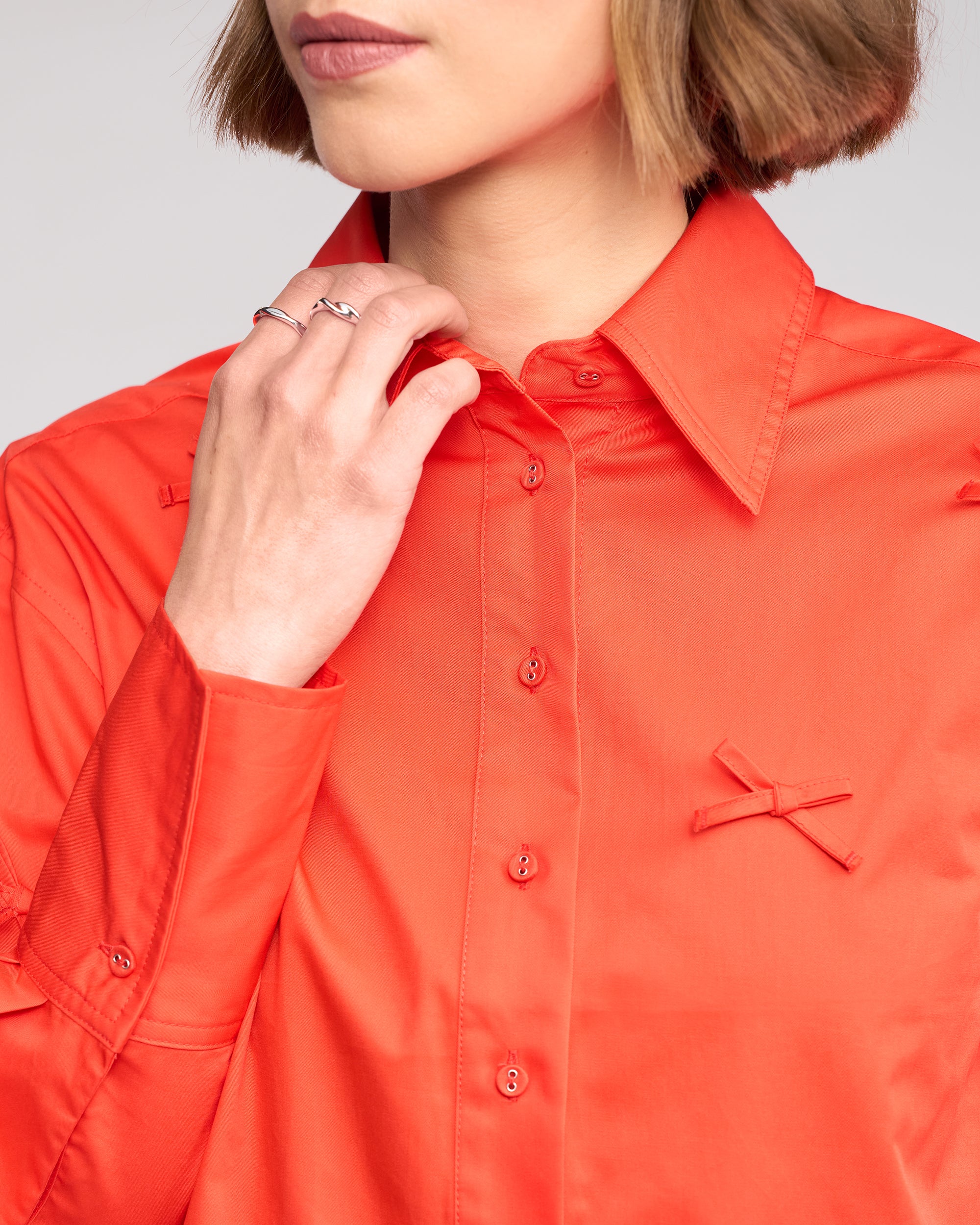 A woman models the Knewe Label Bow Shirt - Lava, a bright orange top featuring a bow appliqué, with her hand touching her collar. Only her lower face and upper torso are shown.