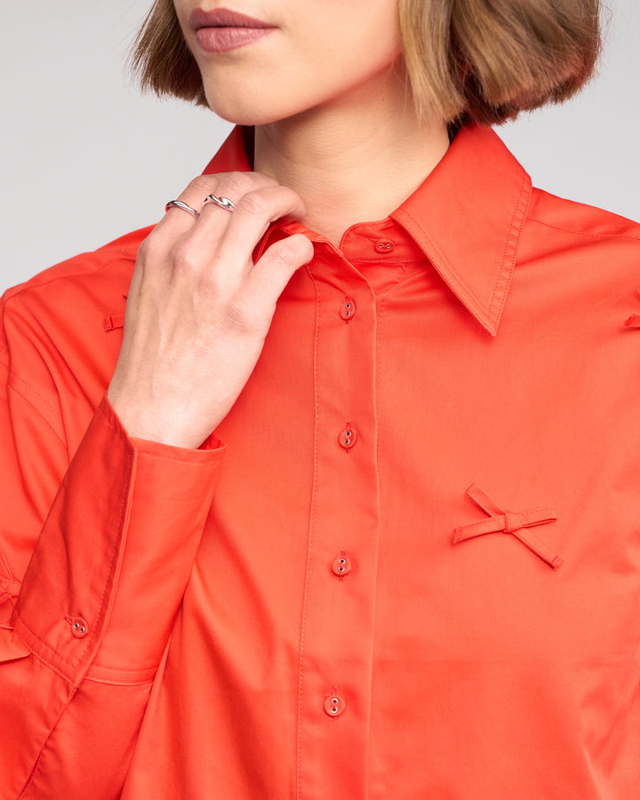 A woman models the Knewe Label Bow Shirt - Lava, a bright orange top featuring a bow appliqué, with her hand touching her collar. Only her lower face and upper torso are shown.
