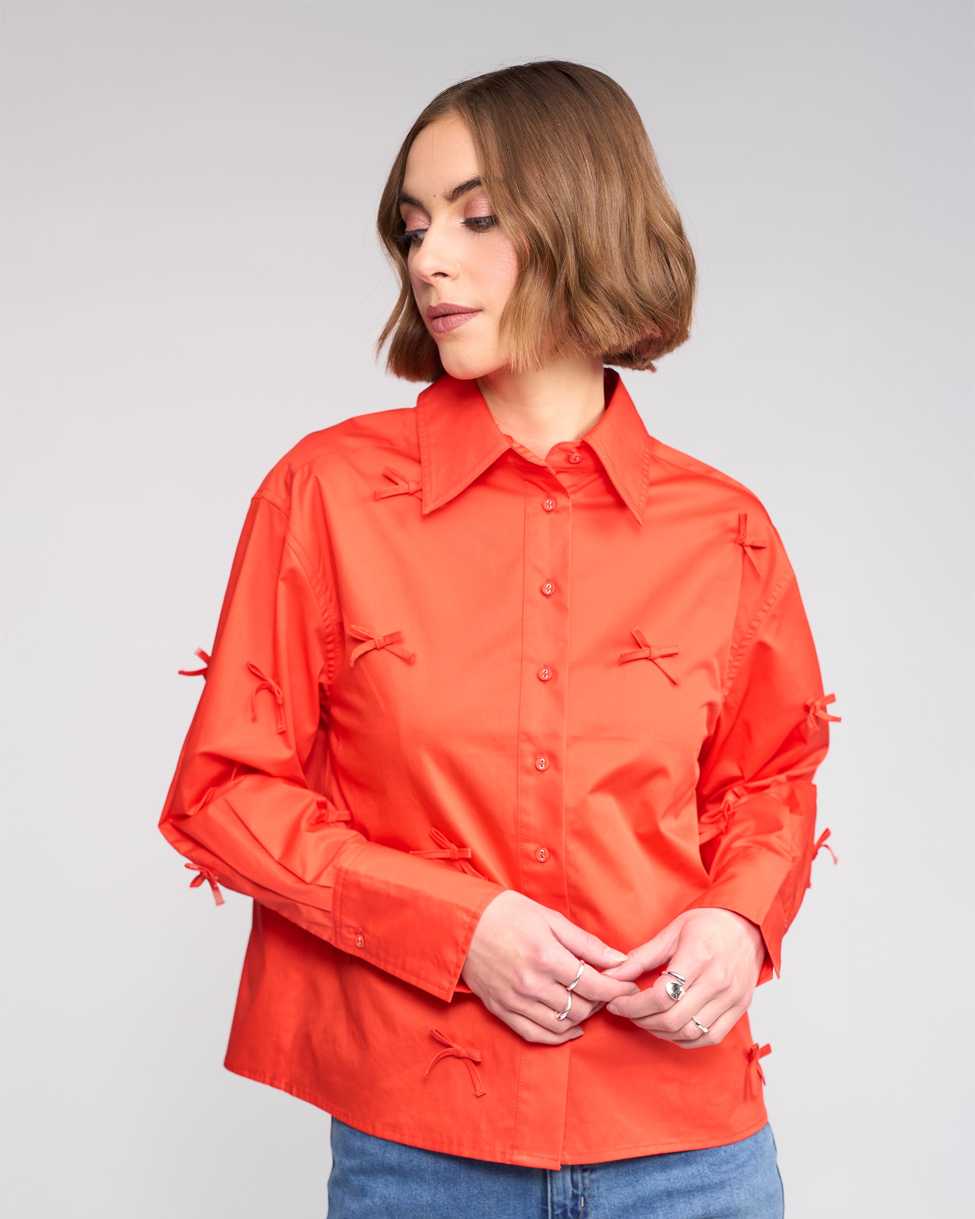 A woman with short brown hair wears the Knewe Label Bow Shirt - Lava with blue jeans. She looks down and to the side, hands gently clasped in front of her, against a plain light-colored background.