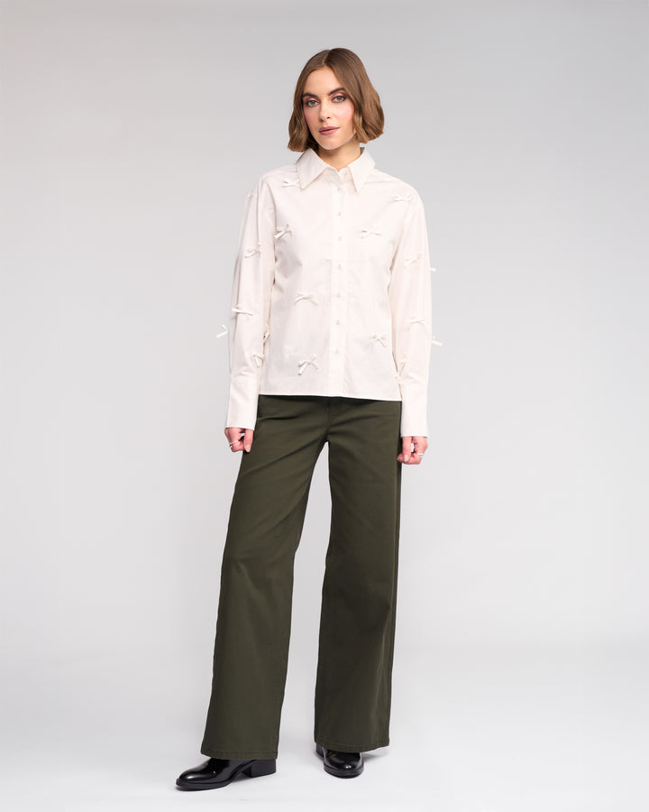 A woman with shoulder-length light brown hair and a neutral expression wears the Knewe Label Bow Shirt - Off White against a plain background. She pairs it with olive wide-leg pants and black shoes.