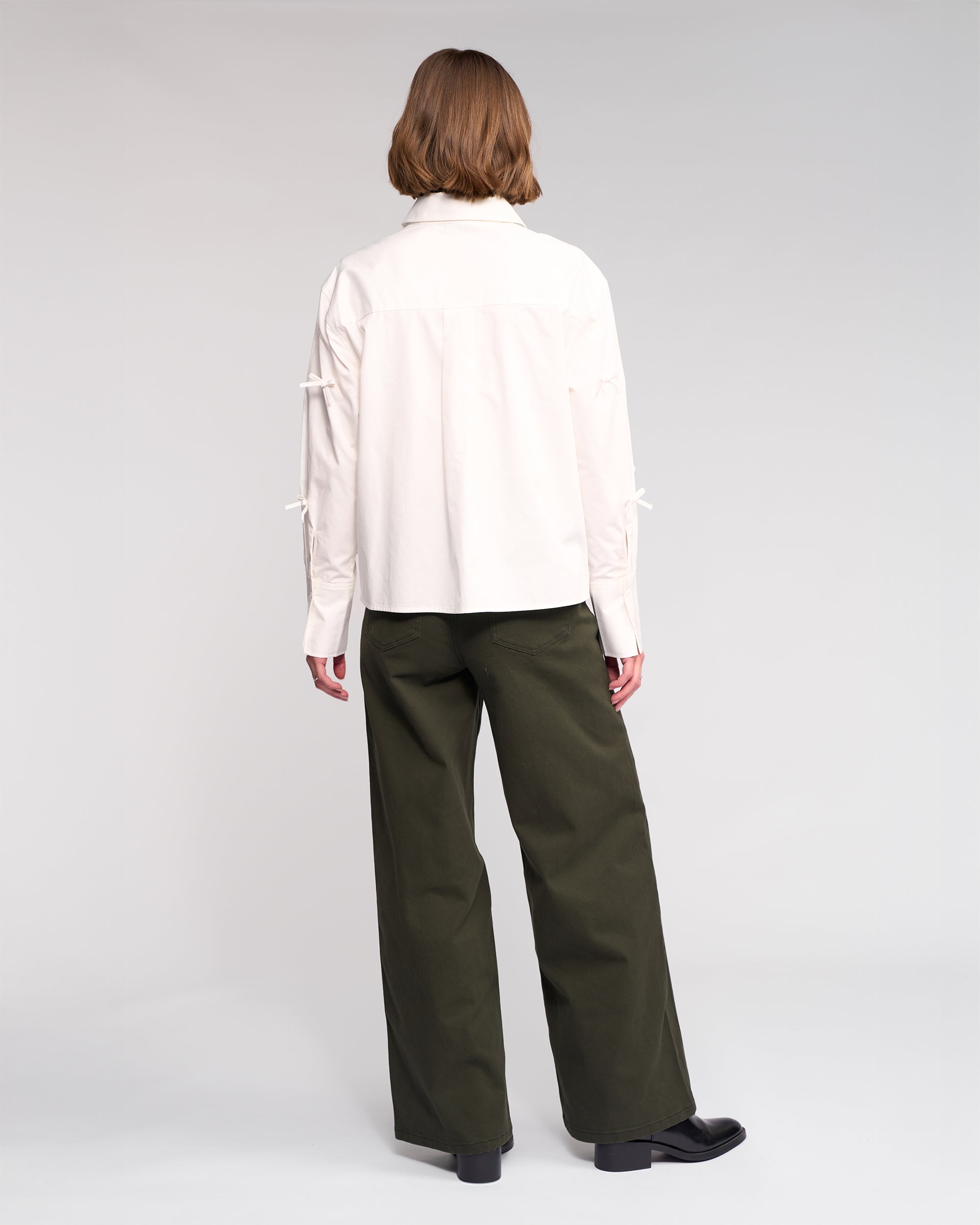 A person with short light brown hair, seen from behind, wears the Knewe Label Bow Shirt in off-white and loose olive-green pants, standing against a plain light background.