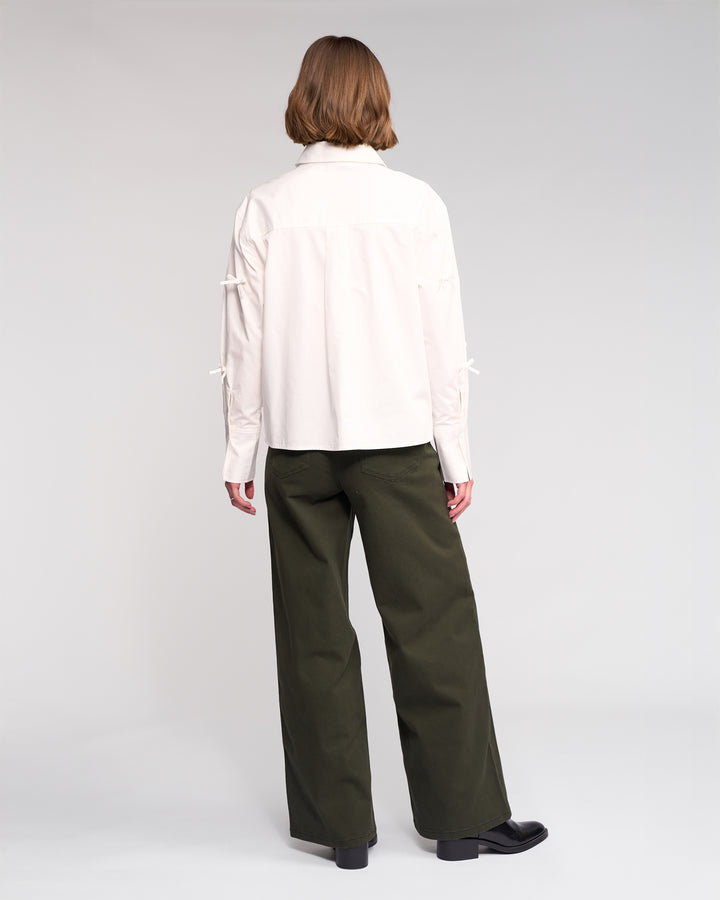 A person with short light brown hair, seen from behind, wears the Knewe Label Bow Shirt in off-white and loose olive-green pants, standing against a plain light background.