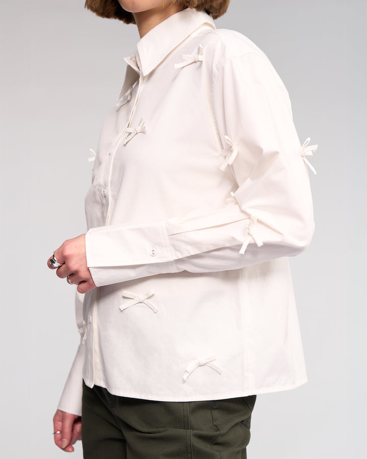 A person wears the Knewe Label Bow Shirt - Off White, featuring small fabric bows, paired with dark green pants. The image is cropped at the chin to highlight the shirt’s delicate details.