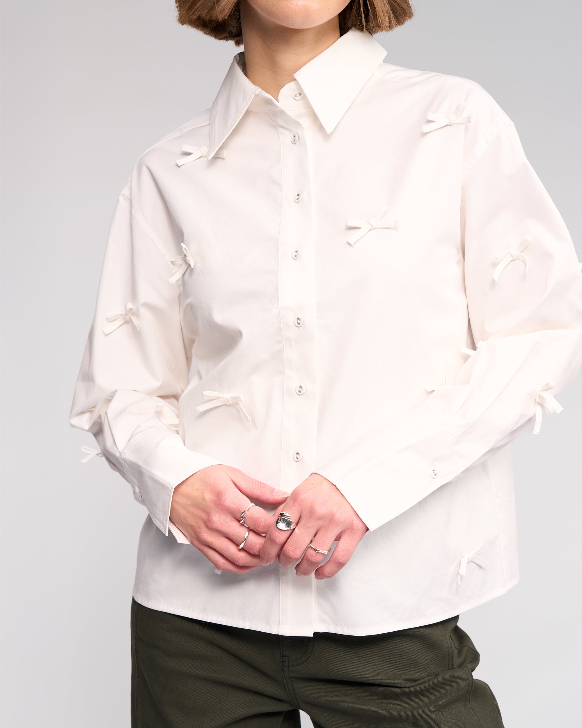 A person wears the Knewe Label Bow Shirt in off white, featuring fabric bow appliqué, paired with olive green pants. Hands are clasped, showcasing multiple rings. The backdrop is plain and light-colored.