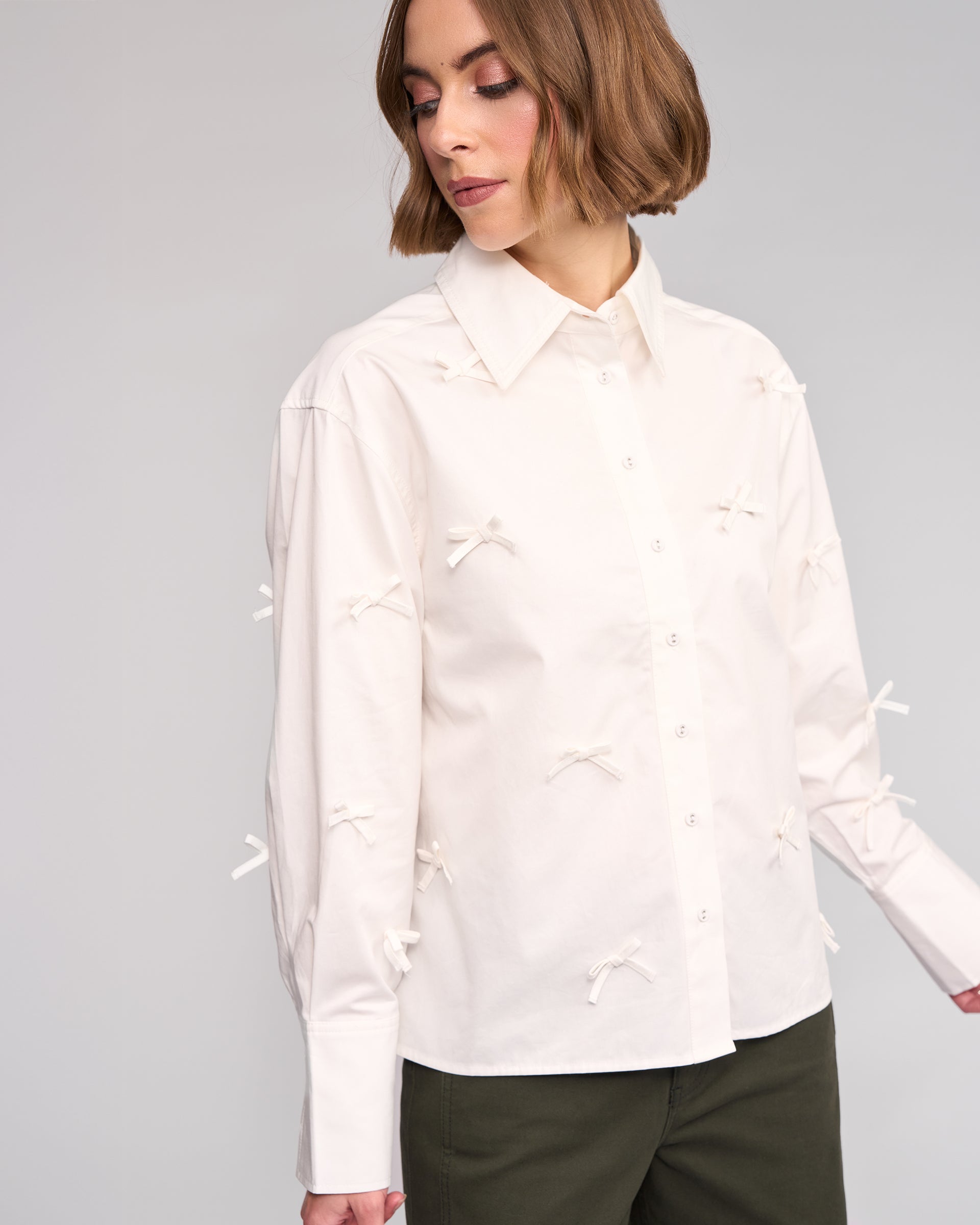 A woman wears the Knewe Label Bow Shirt in off white, featuring delicate bow appliqué, paired with dark green pants against a light gray background.