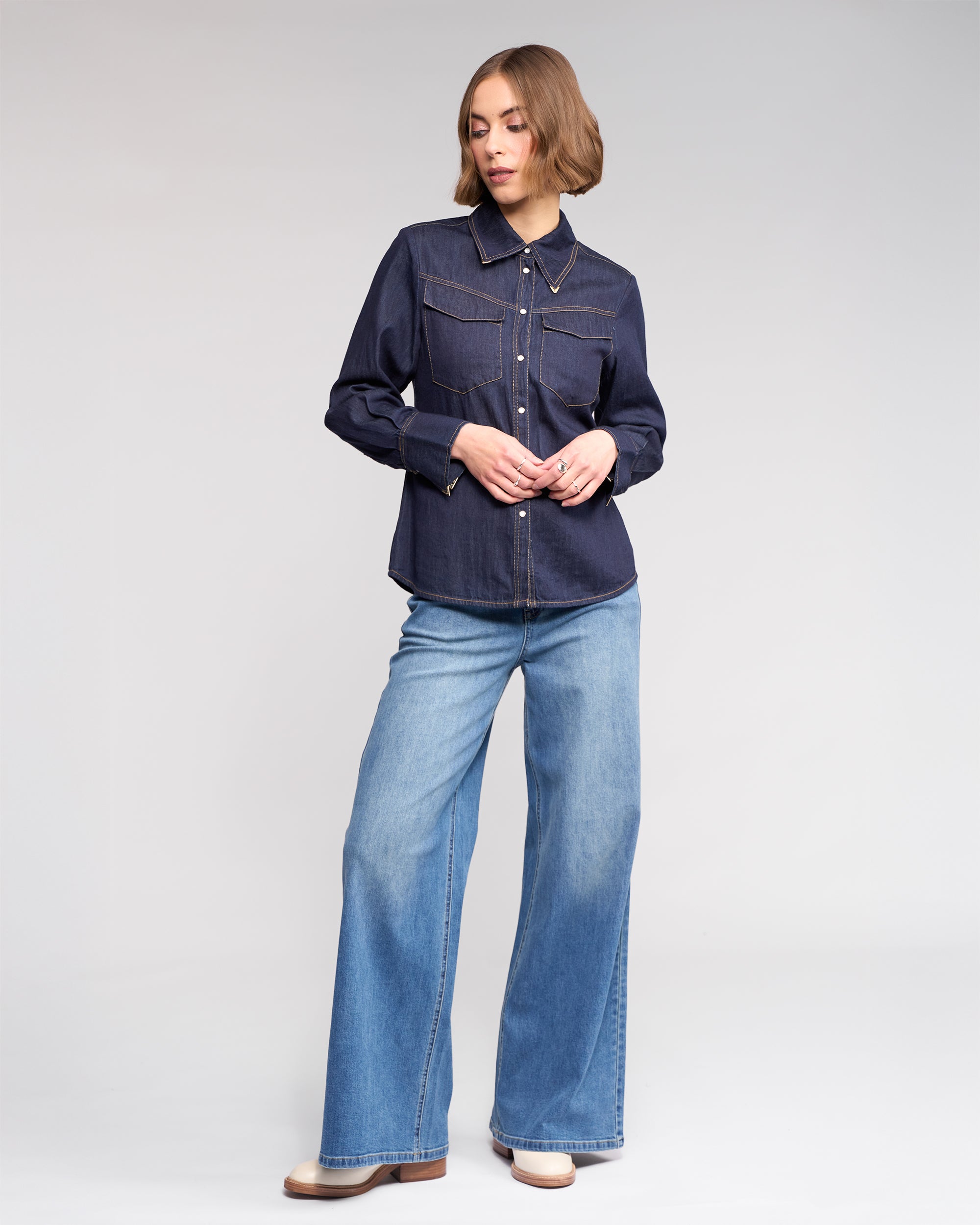A woman with short, wavy hair stands against a plain background wearing the Knewe Label Nash Shirt in Dark Denim, featuring western-inspired details and front pockets, paired with light wide-leg jeans. She looks to the side, hand gently touching her fingers.