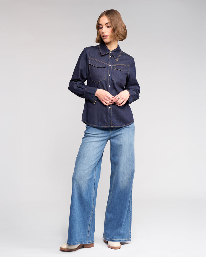 A woman with short, wavy hair stands against a plain background wearing the Knewe Label Nash Shirt in Dark Denim, featuring western-inspired details and front pockets, paired with light wide-leg jeans. She looks to the side, hand gently touching her fingers.