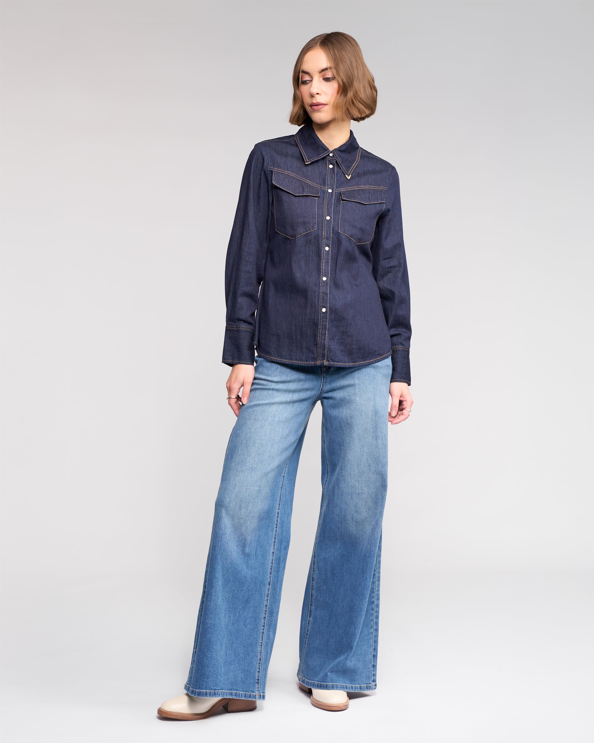 A woman wears the Knewe Label Nash Shirt in dark denim, styled with light blue wide-leg jeans and tan boots, standing against a plain light background.