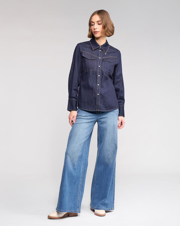 A woman wears the Knewe Label Nash Shirt in dark denim, styled with light blue wide-leg jeans and tan boots, standing against a plain light background.