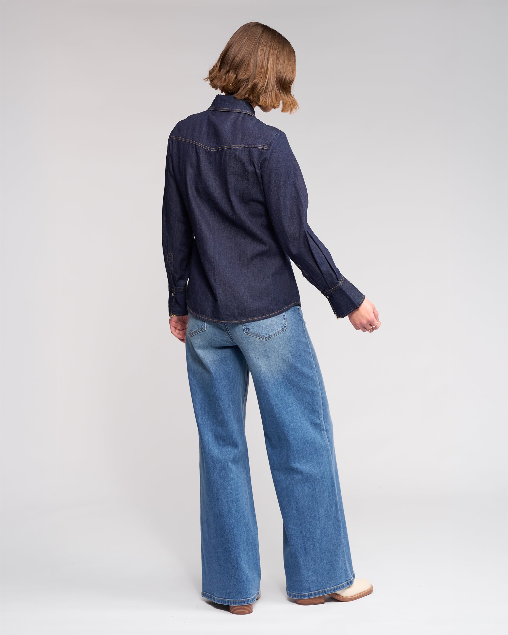A person with short brown hair faces away, wearing the Knewe Label Nash Shirt in dark denim with western-inspired details and light blue wide-leg jeans, set against a plain light background.