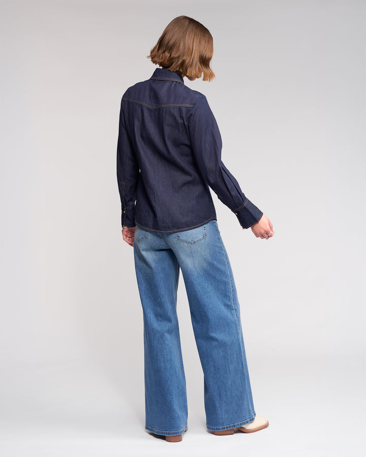 A person with short brown hair faces away, wearing the Knewe Label Nash Shirt in dark denim with western-inspired details and light blue wide-leg jeans, set against a plain light background.