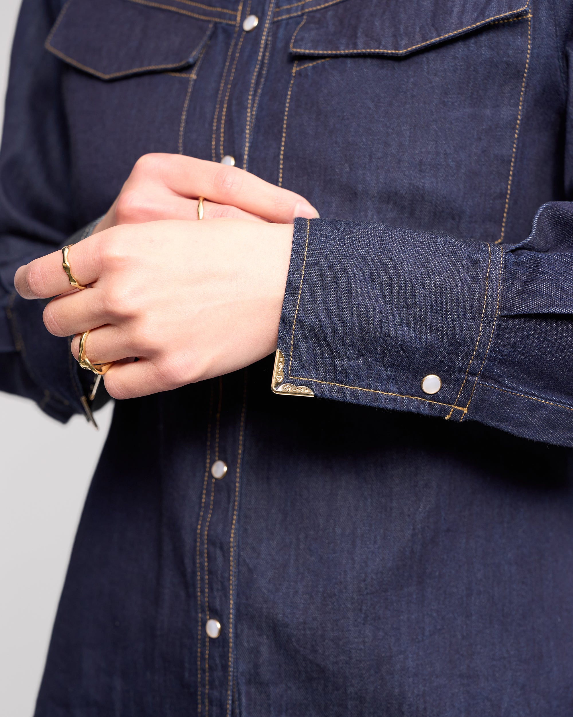 Close-up of a person wearing the Knewe Label Nash Shirt in Dark Denim, featuring western-inspired white snaps and yellow stitching. Their hands are folded, displaying several gold rings on their fingers.