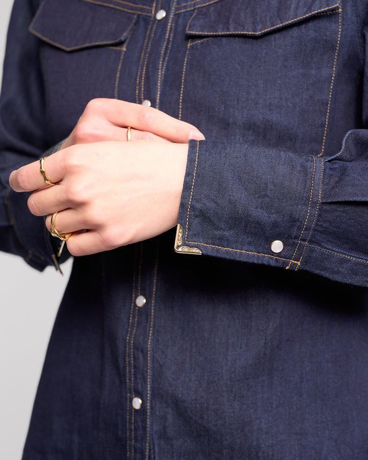 Close-up of a person wearing the Knewe Label Nash Shirt in Dark Denim, featuring western-inspired white snaps and yellow stitching. Their hands are folded, displaying several gold rings on their fingers.