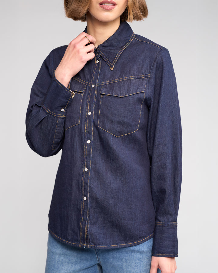 A woman with short brown hair wears the Knewe Label Nash Shirt in Dark Denim, featuring western-inspired details and two chest pockets. She pairs it with light blue jeans and touches her collar against a plain light gray background.