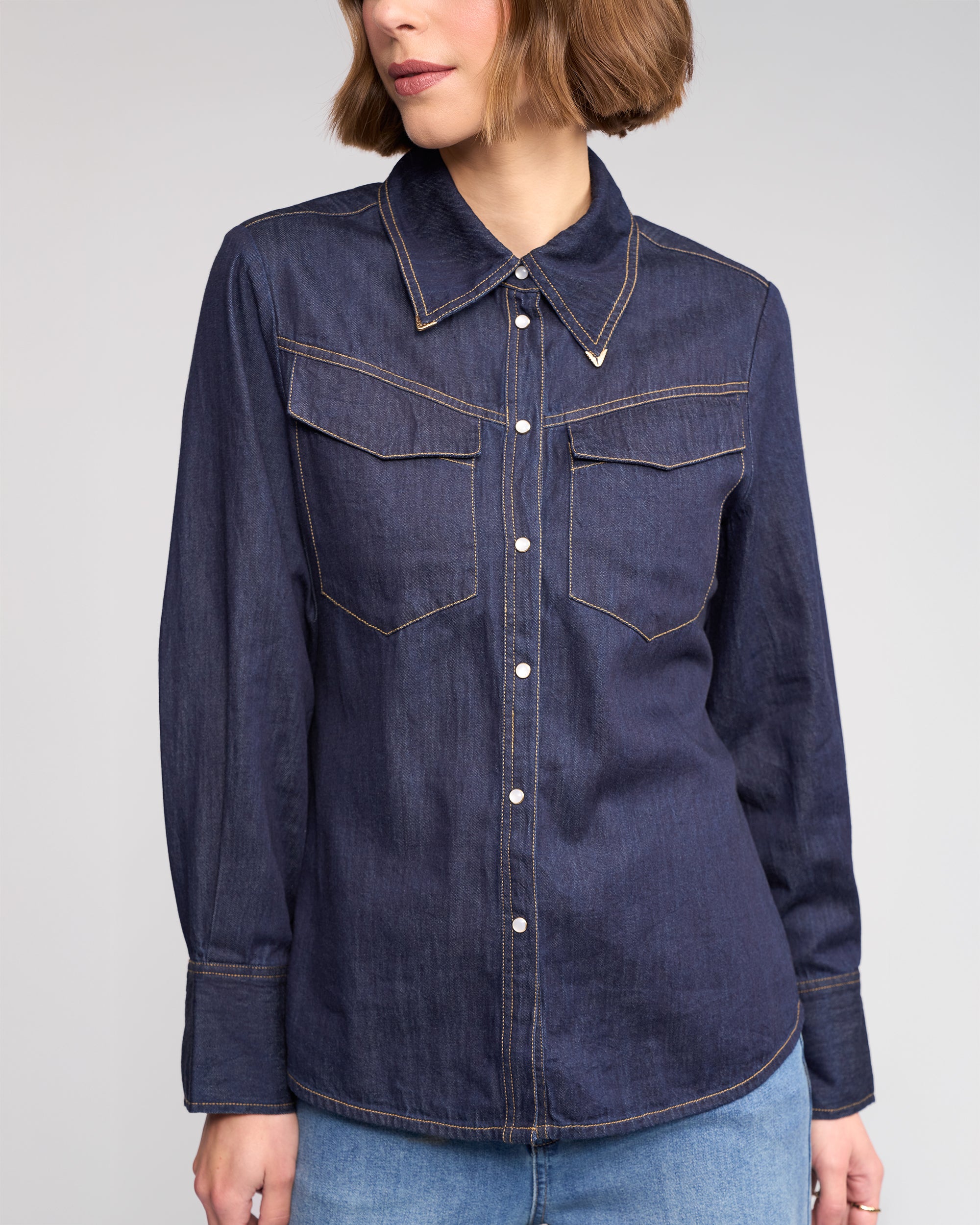 A woman wears the Nash Shirt in Dark Denim by Knewe Label, featuring western-inspired details and two chest pockets, paired with light blue jeans. She stands against a plain background with her face partially out of frame.