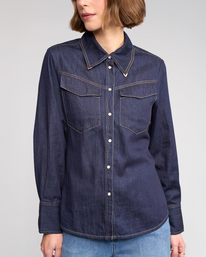 A woman wears the Nash Shirt in Dark Denim by Knewe Label, featuring western-inspired details and two chest pockets, paired with light blue jeans. She stands against a plain background with her face partially out of frame.