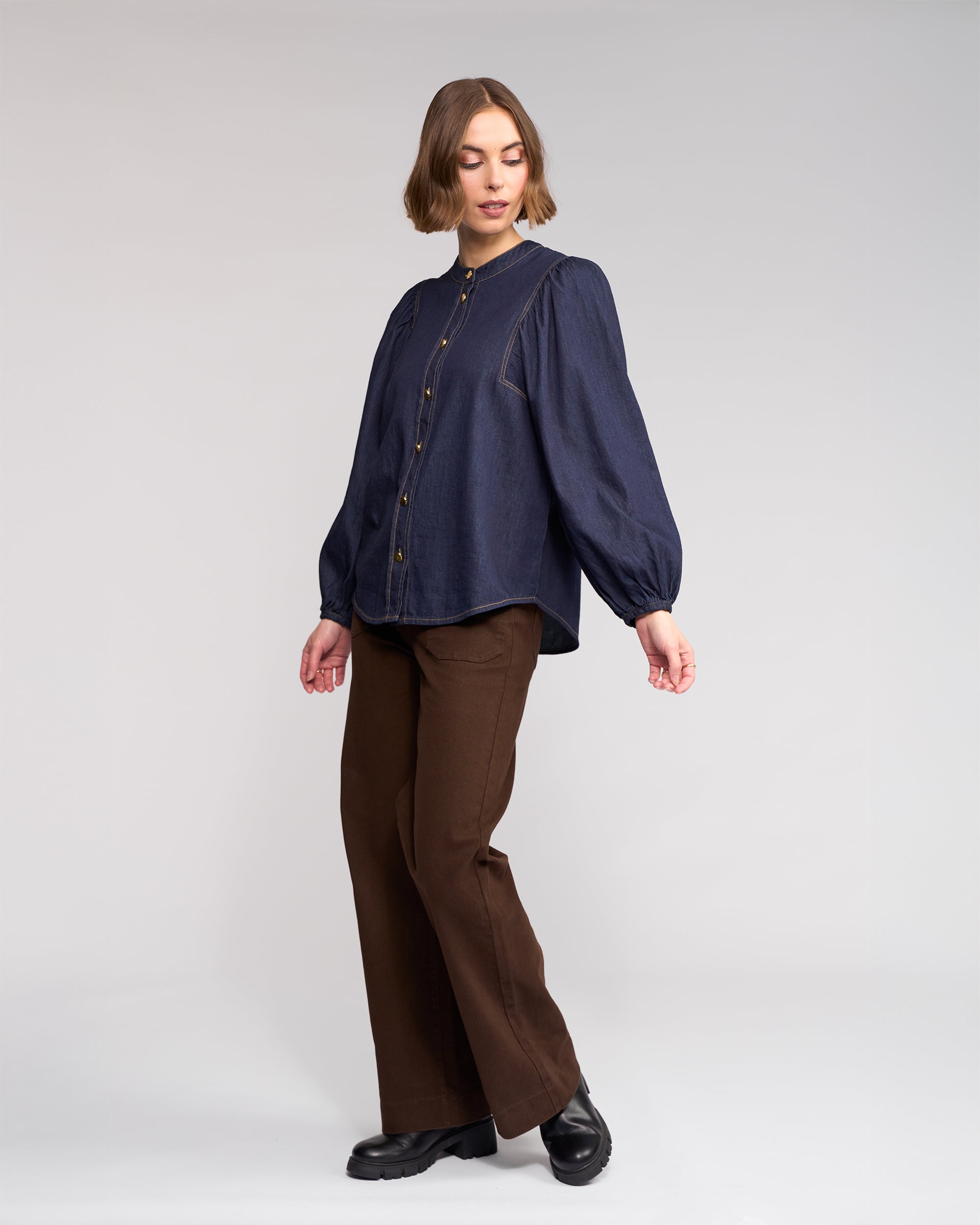 A person with short hair stands against a plain background, wearing the Knewe Label Jesse Shirt in Dark Denim with puffed sleeves and gold buttons, brown wide-leg pants, and black shoes, looking slightly to the side.