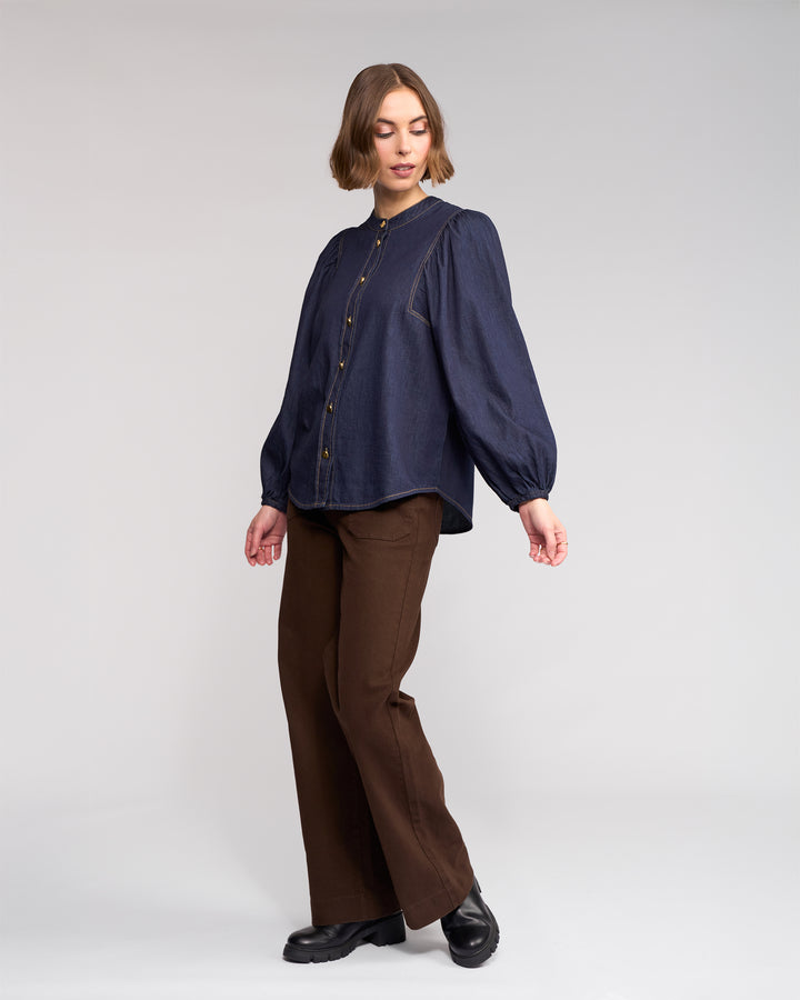 A person with short hair stands against a plain background, wearing the Knewe Label Jesse Shirt in Dark Denim with puffed sleeves and gold buttons, brown wide-leg pants, and black shoes, looking slightly to the side.