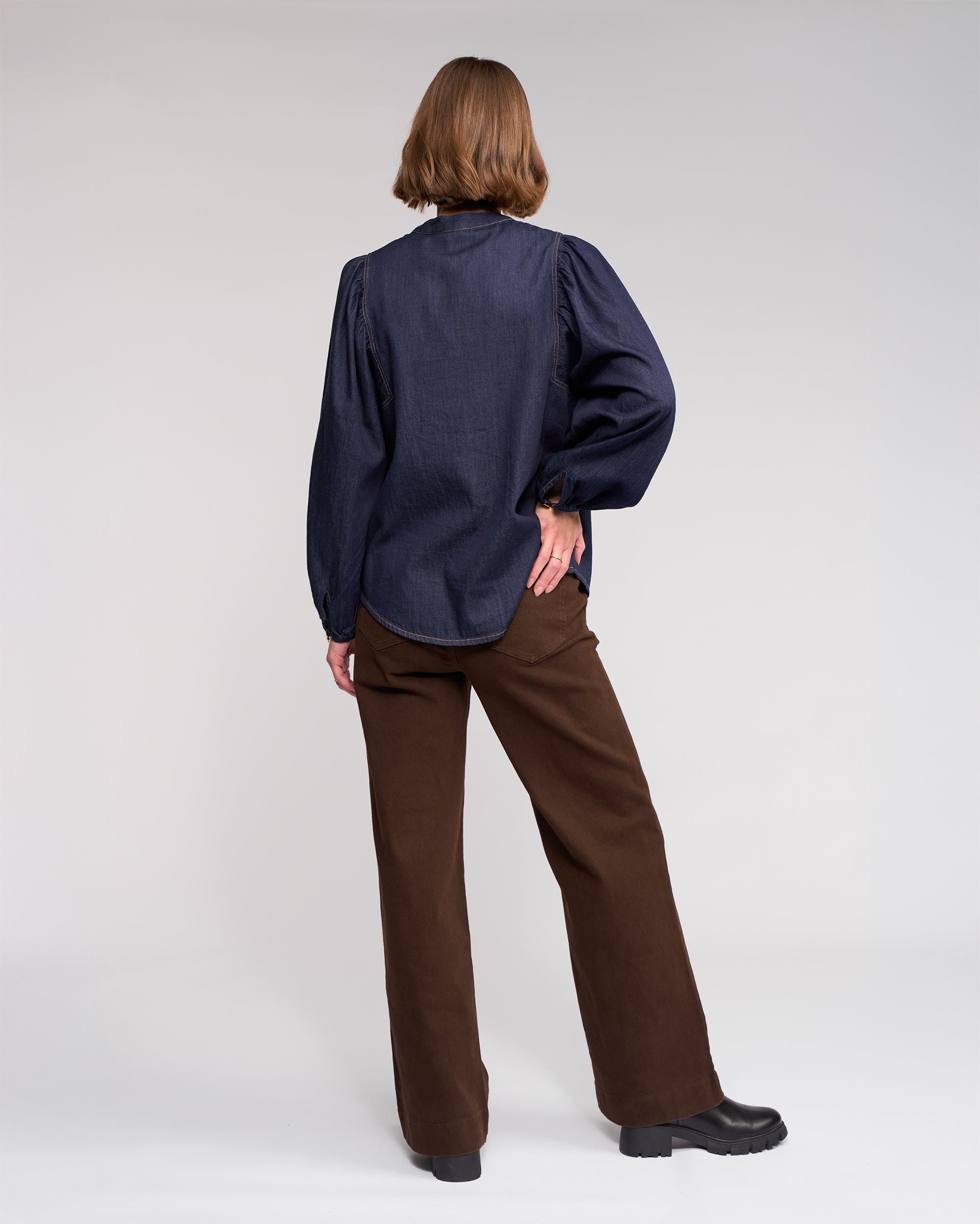A person stands with their back to the camera, wearing the Jesse Shirt - Dark Denim by Knewe Label, paired with brown wide-leg pants and black shoes, set against a plain light background.