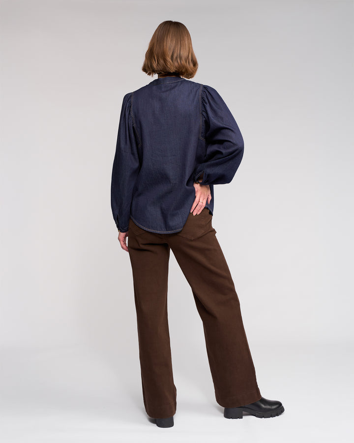 A person stands with their back to the camera, wearing the Jesse Shirt - Dark Denim by Knewe Label, paired with brown wide-leg pants and black shoes, set against a plain light background.
