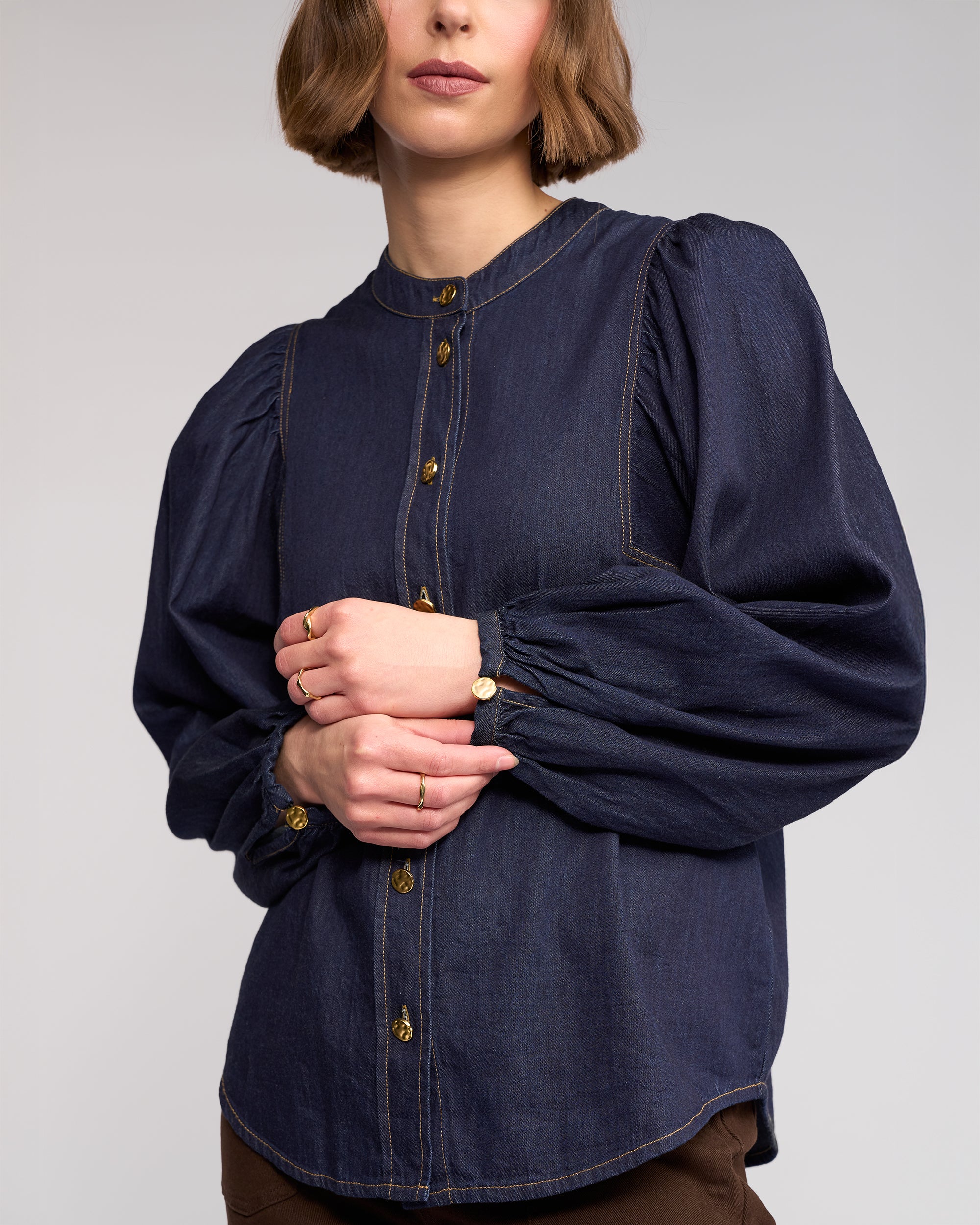 A woman with short brown hair models the Knewe Label Jesse Shirt in Dark Denim, featuring puffed sleeves and gold buttons, paired with gold rings and a watch, standing against a plain light background.