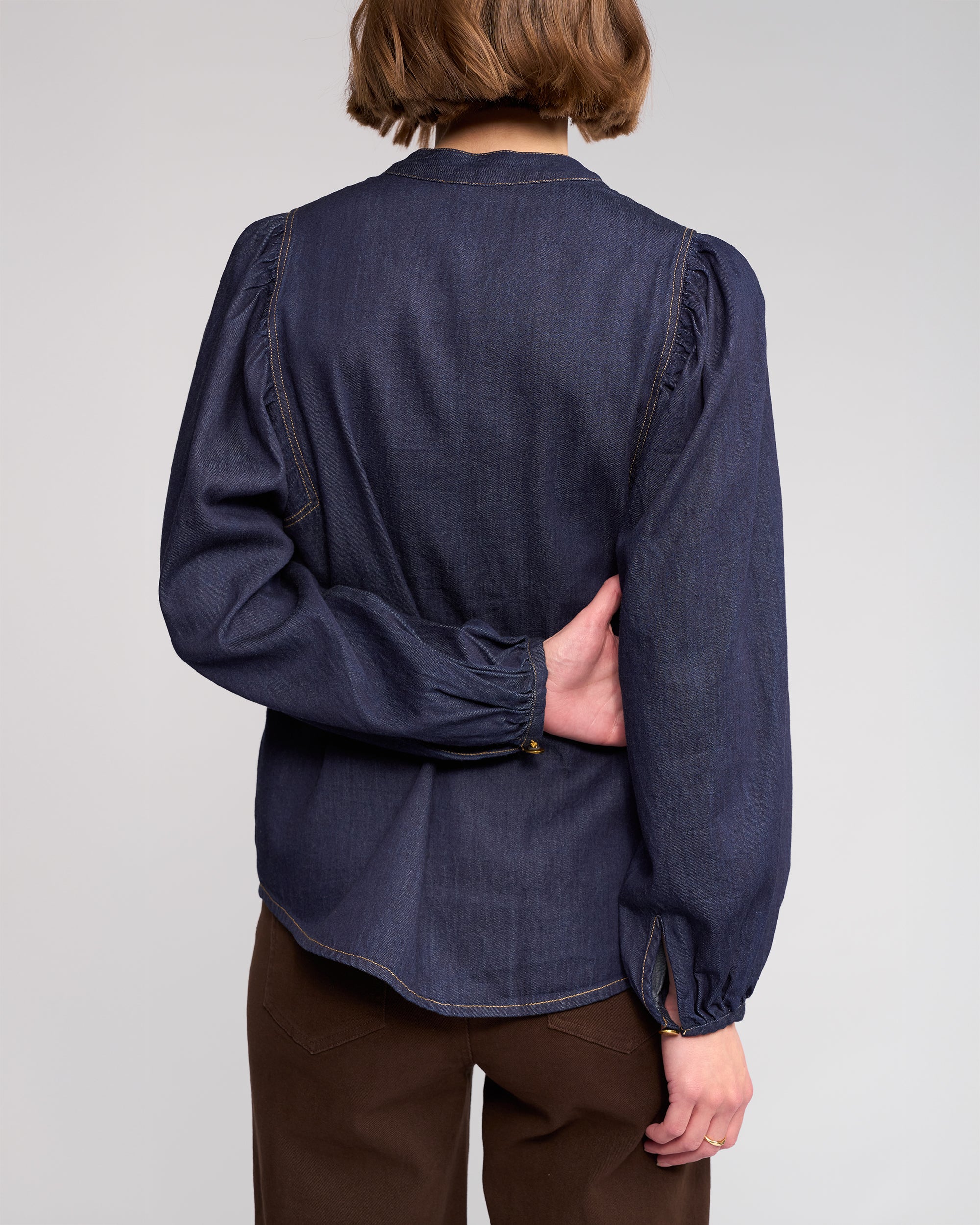 A person with short brown hair is shown from the back, wearing the Knewe Label Jesse Shirt in dark denim and brown pants, left arm bent behind them holding their right side.