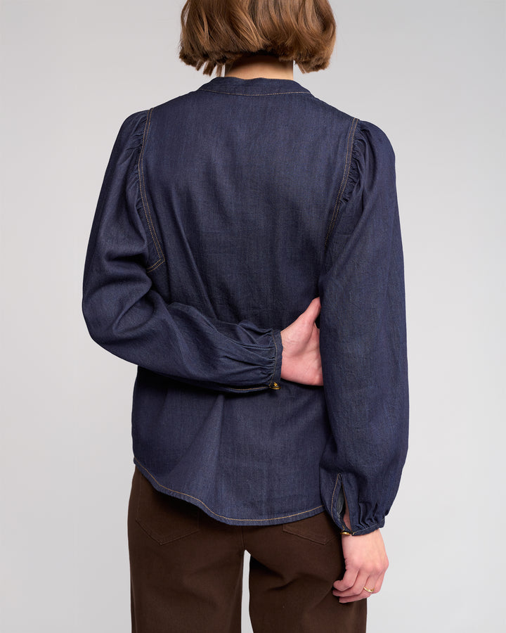 A person with short brown hair is shown from the back, wearing the Knewe Label Jesse Shirt in dark denim and brown pants, left arm bent behind them holding their right side.