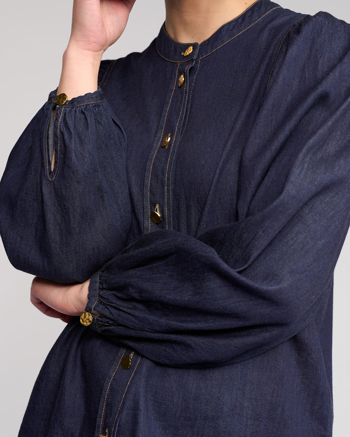 A person in the Knewe Label Jesse Shirt – Dark Denim, featuring gold buttons and long sleeves, stands with one arm crossed and a hand touching their face. Their head is partially out of frame.