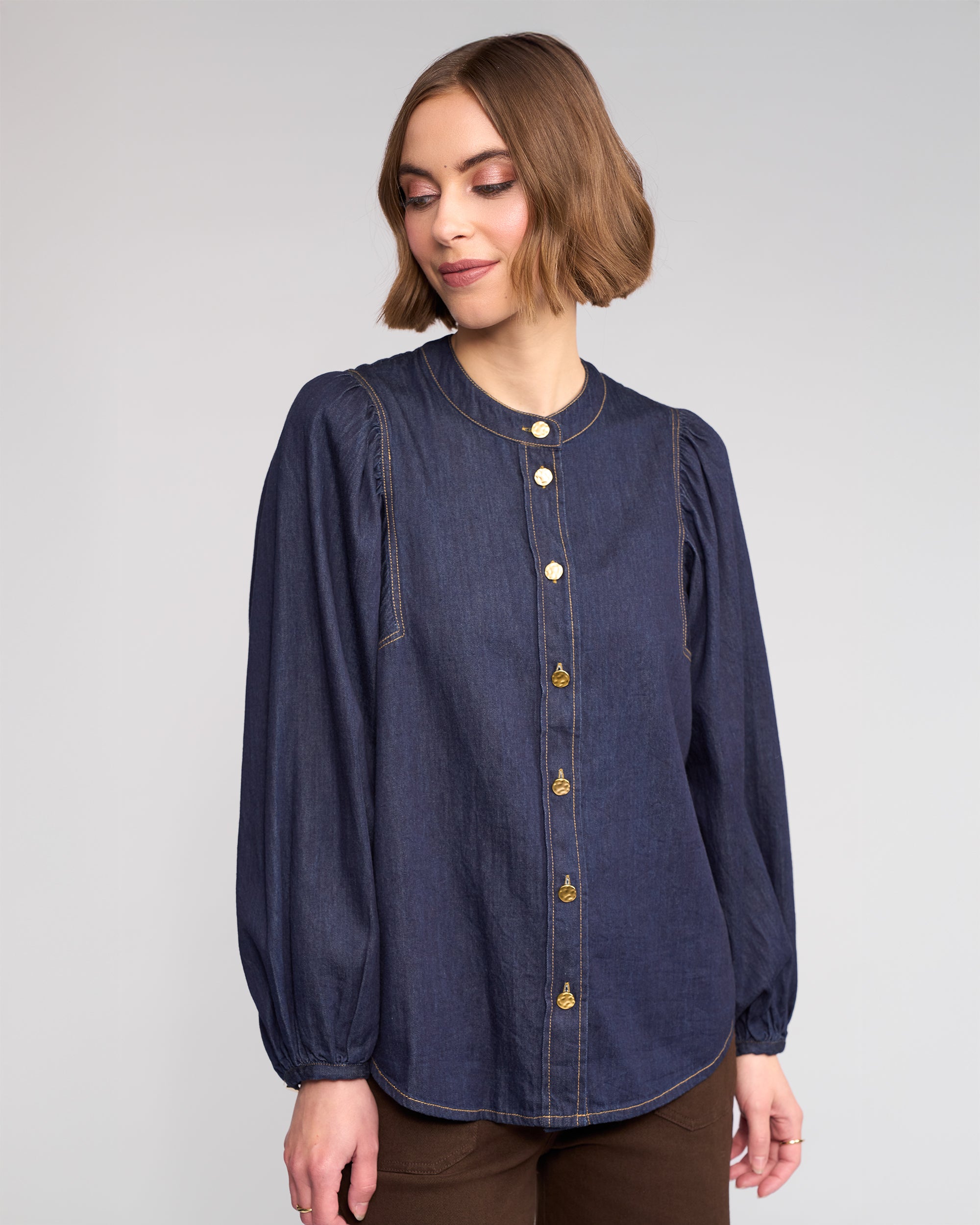 A woman with a short bob wears Knewe Label’s Jesse Shirt in dark denim—collarless, with puffed sleeves and gold buttons—paired with brown pants. She looks slightly down against a plain background.