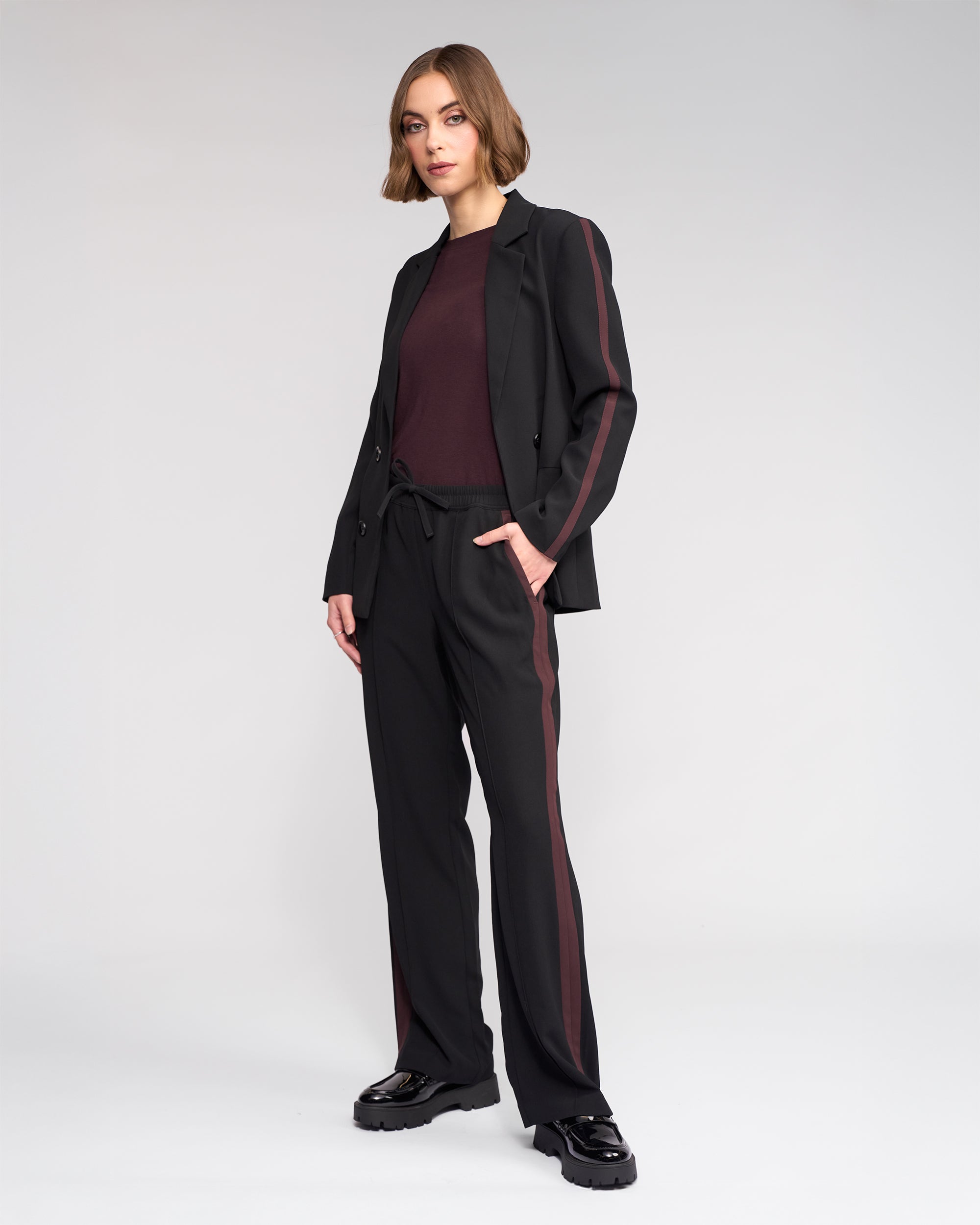 A woman with a bob haircut and neutral expression stands against a light gray background wearing the Knewe Label Clementine Blazer in Black/Raisin, featuring contrast sleeves and maroon stripes, paired with a maroon top and black chunky shoes.