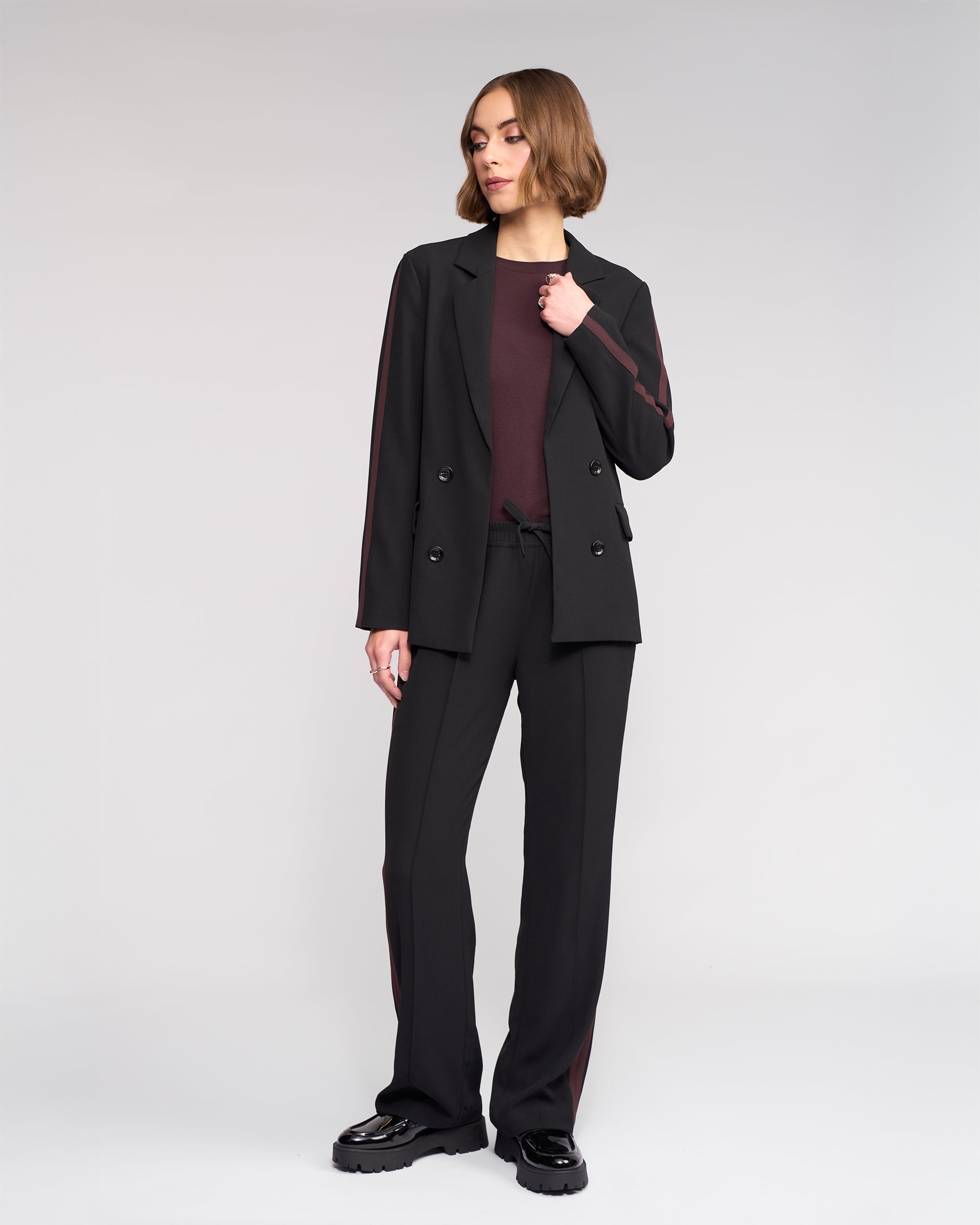 A woman with short brown hair models the Knewe Label Clementine Blazer in Black/Raisin, standing against a light background with one hand by her side and the other relaxed, looking slightly to her left.