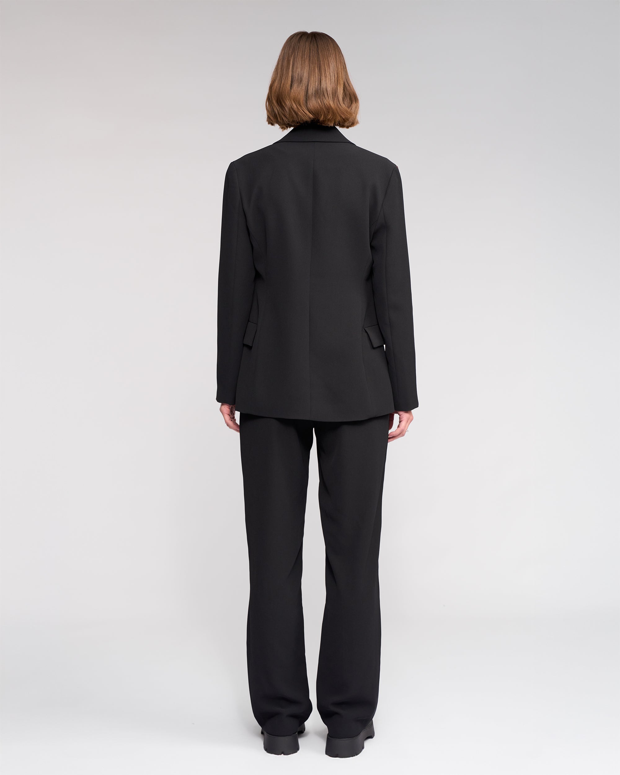 Seen from behind, a person with short, light brown hair wears the Knewe Label Clementine Blazer in Black/Raisin with contrast sleeve detailing, paired with black trousers, against a plain light background.