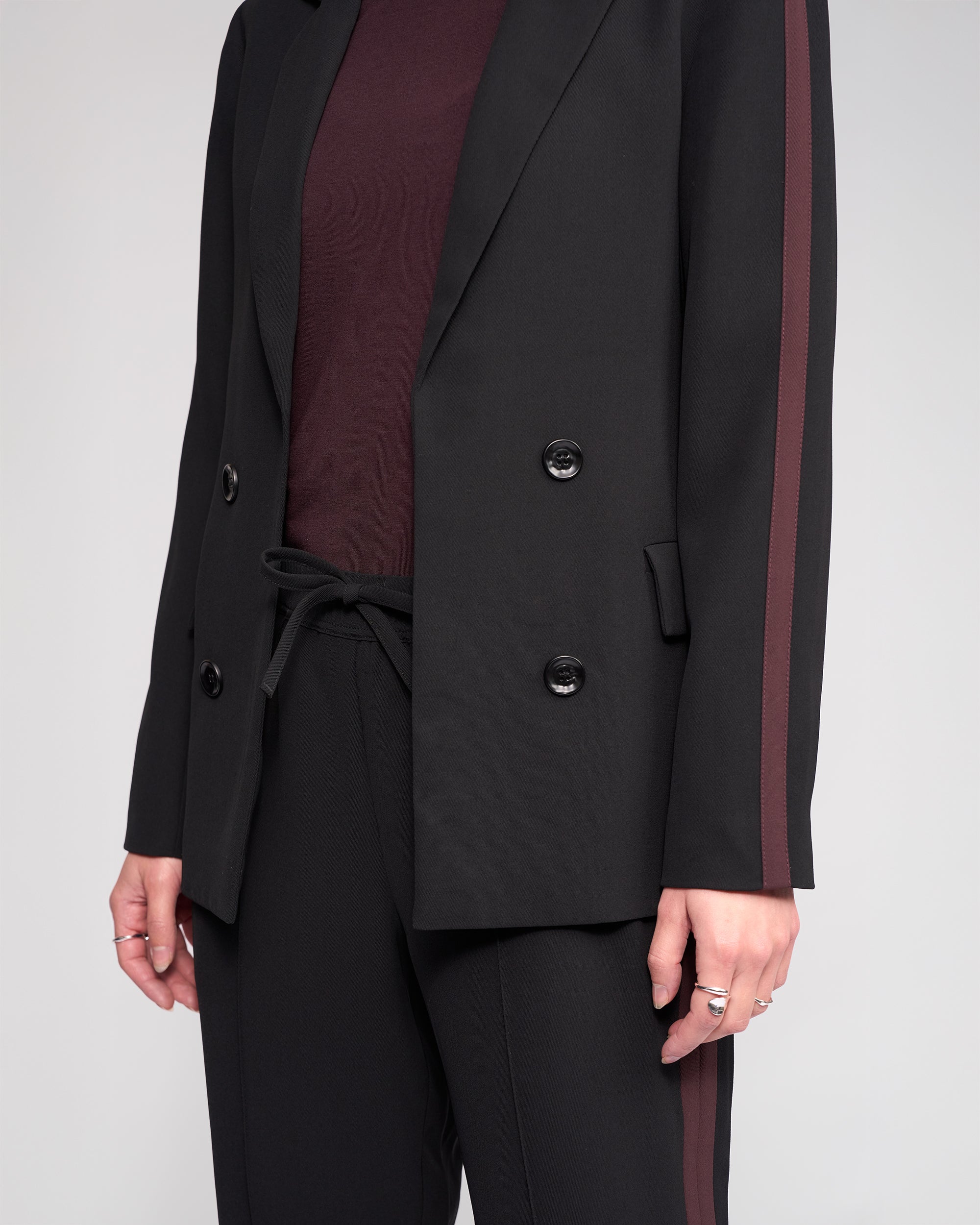 A person wears the Knewe Label Clementine Blazer in Black/Raisin with maroon contrast sleeves and side stripes, paired with a matching maroon top and drawstring pants; the double-breasted blazer and rings complete the chic look.