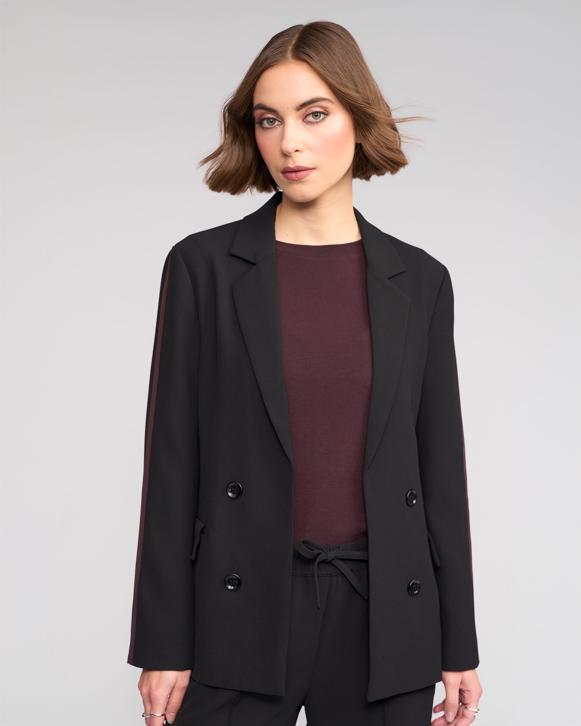 A woman with short brown hair wears the Knewe Label Clementine Blazer in Black/Raisin over a dark top and matching black pants. She stands against a light gray background, looking confidently at the camera.