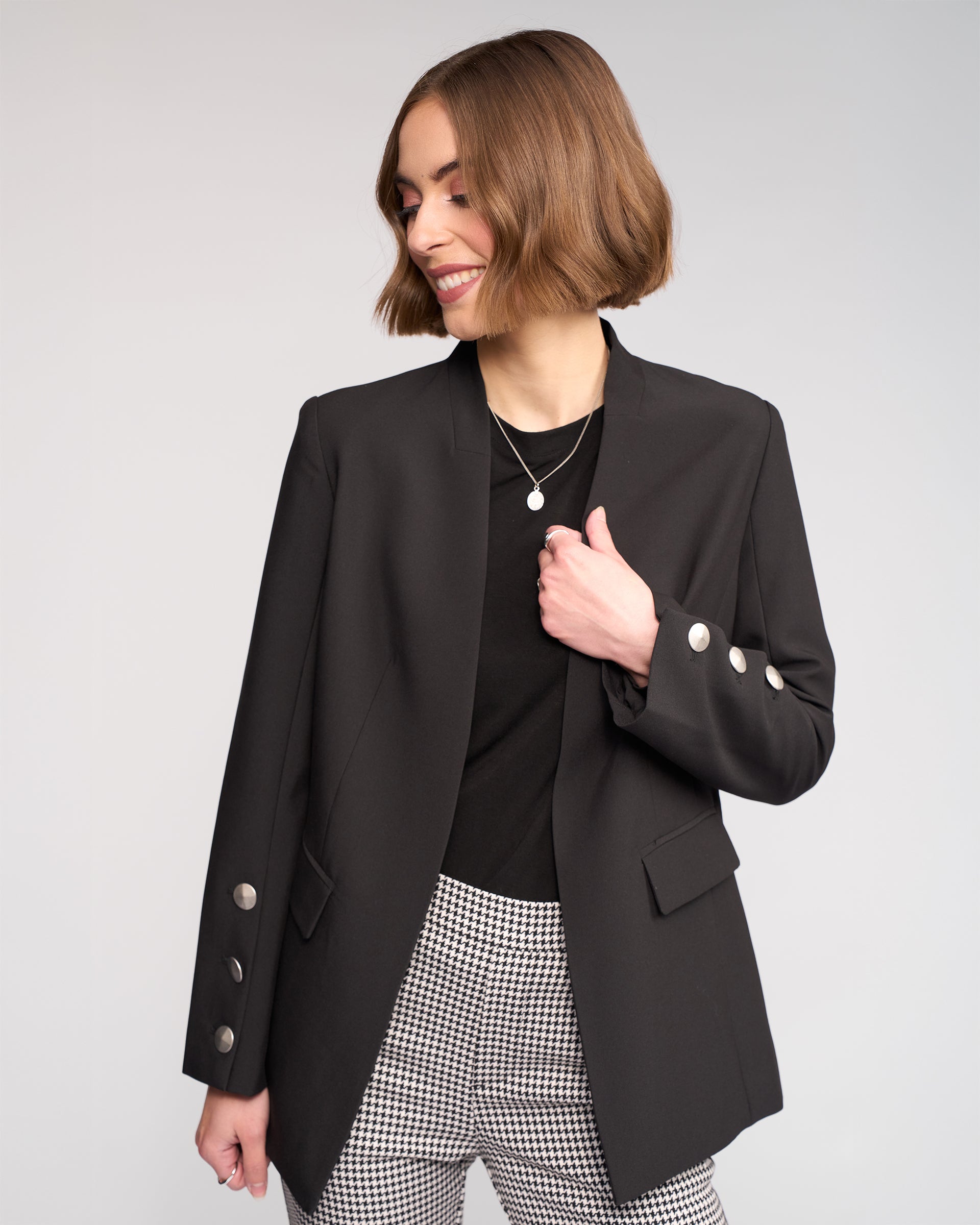 A woman with a chin-length bob smiles while looking to the side, wearing Knewe Label’s Carter Blazer in black with silver buttons over a black top and checkered pants, standing against a plain light background.