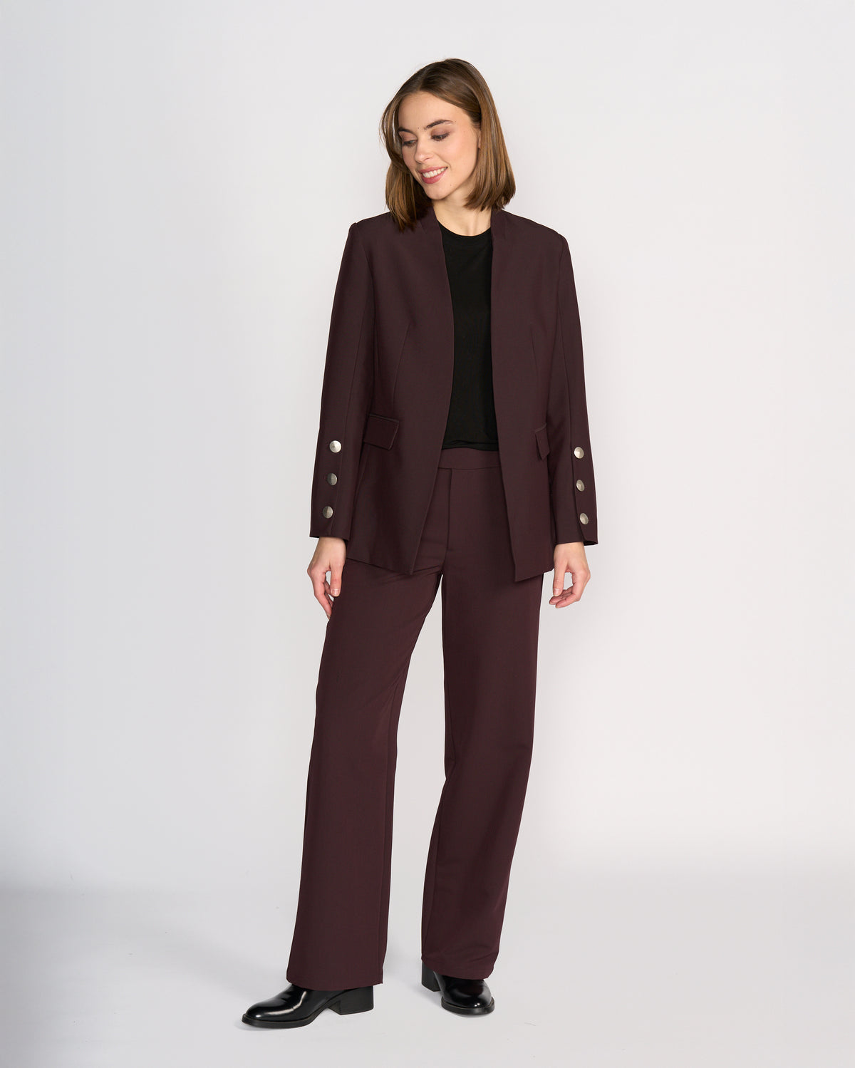 A woman models the Knewe Label Carter Blazer in Raisin against a white background, paired with a black top and shoes. She gazes to the side and smiles, highlighting the blazer's modern silhouette and silver button details on the sleeves.