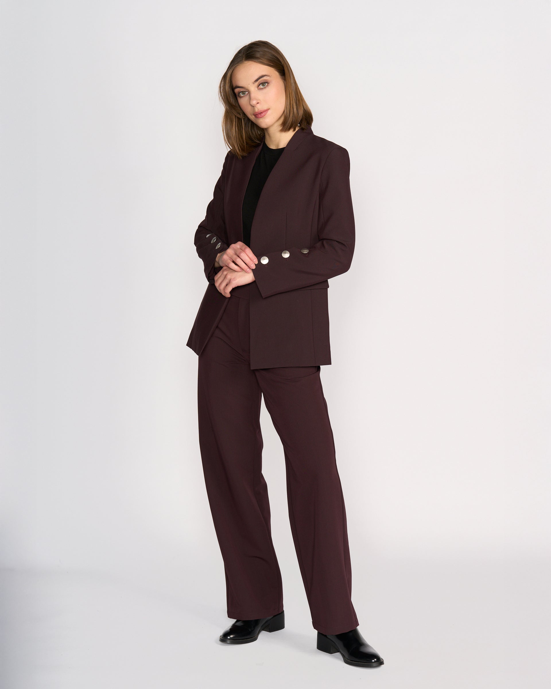 A woman with shoulder-length hair stands against a light background, wearing the Knewe Label Carter Blazer in Raisin with silver buttons, paired with a black top and shoes. She poses with her hands clasped.