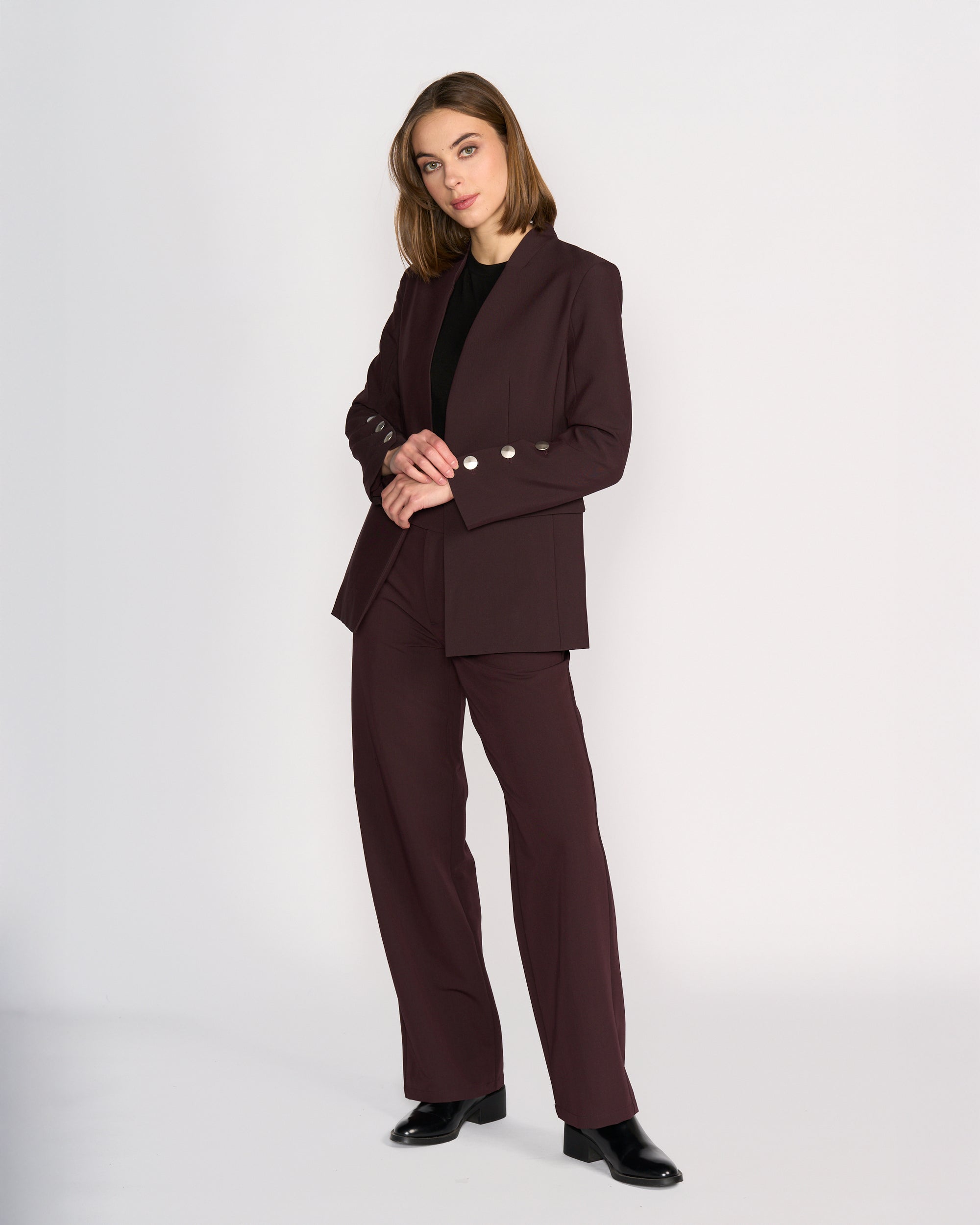 A woman with shoulder-length hair stands against a light background, wearing the Knewe Label Carter Blazer in Raisin with silver buttons, paired with a black top and shoes. She poses with her hands clasped.
