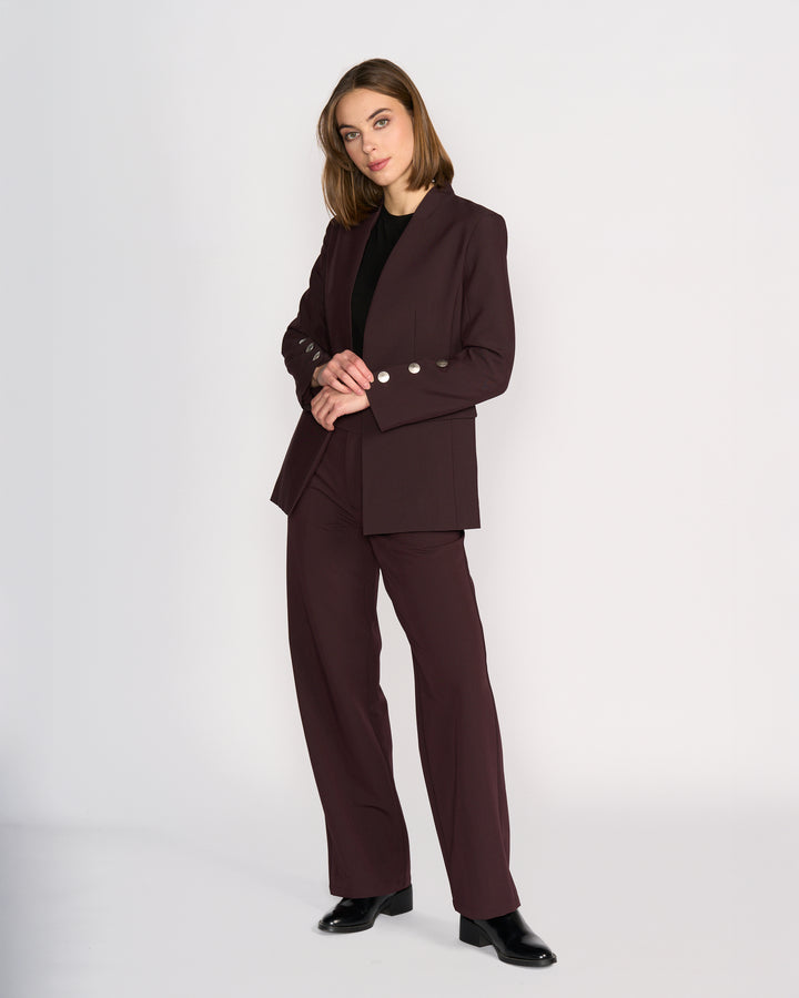 A woman with shoulder-length hair stands against a light background, wearing the Knewe Label Carter Blazer in Raisin with silver buttons, paired with a black top and shoes. She poses with her hands clasped.