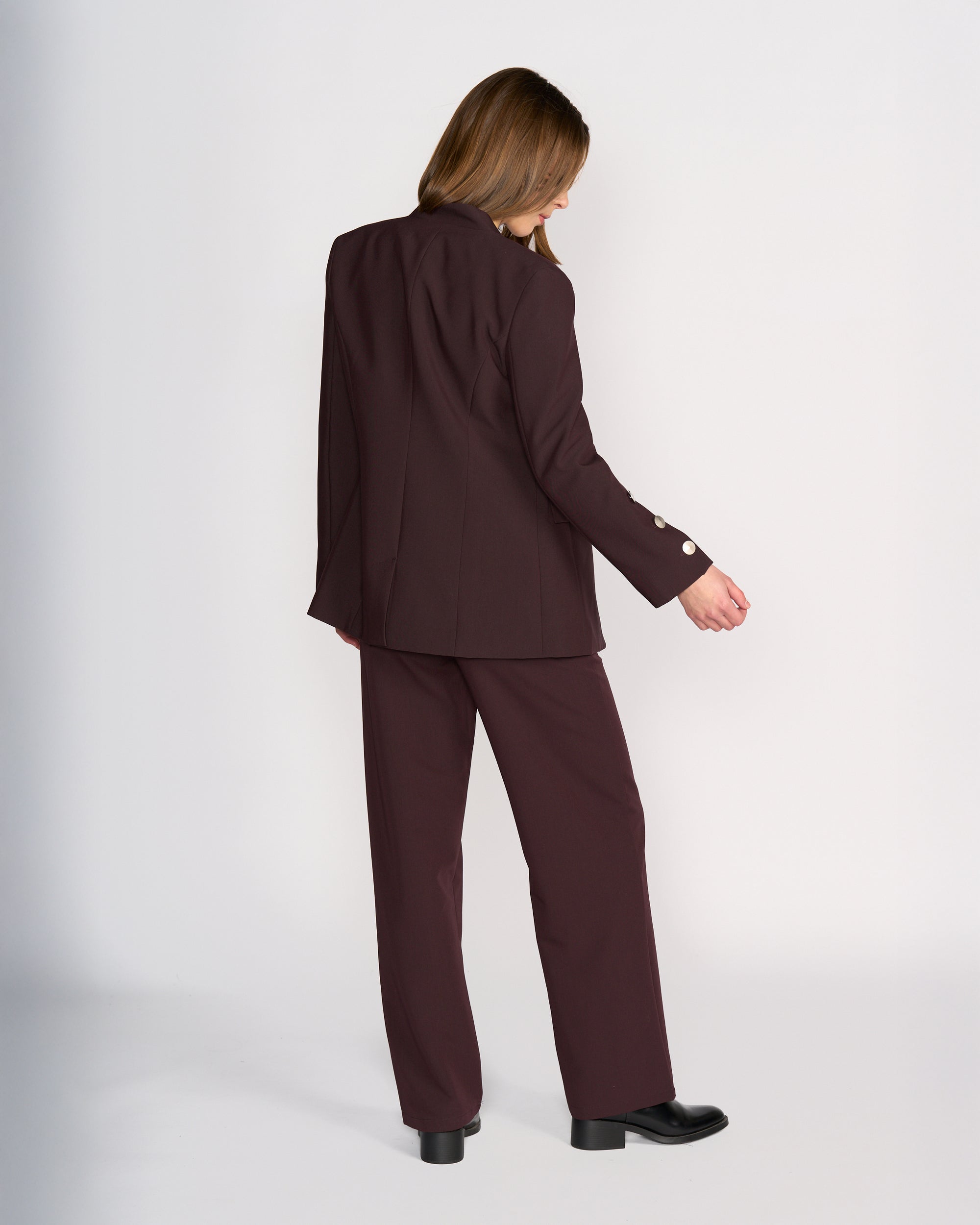A woman with light brown hair stands facing away, wearing the Knewe Label Carter Blazer in Raisin with matching trousers and black shoes, set against a plain light background.