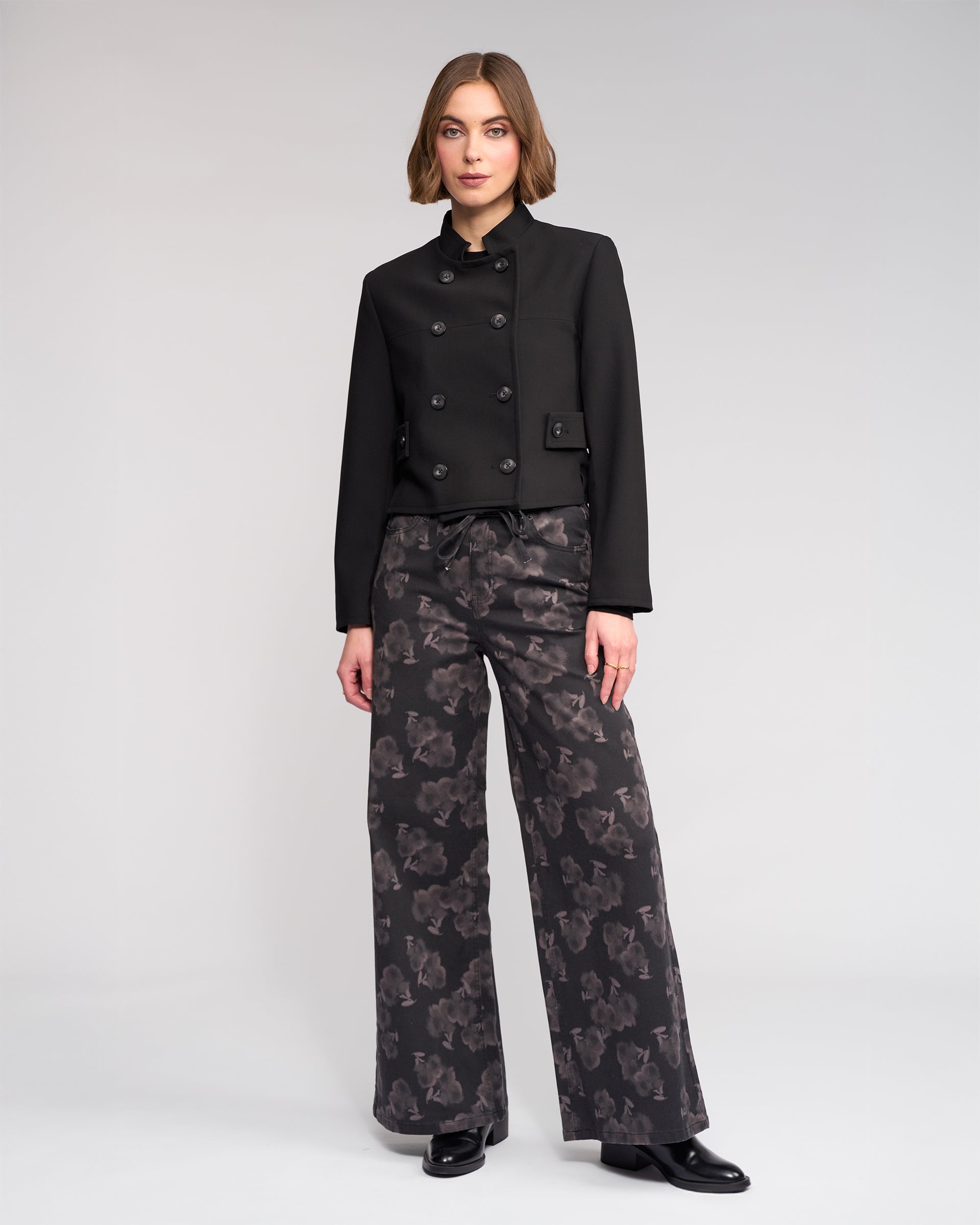A woman with short, light brown hair and a neutral expression stands against a plain background wearing Knewe Label's Soho Jacket in black, paired with wide-leg patterned black trousers and black shoes.