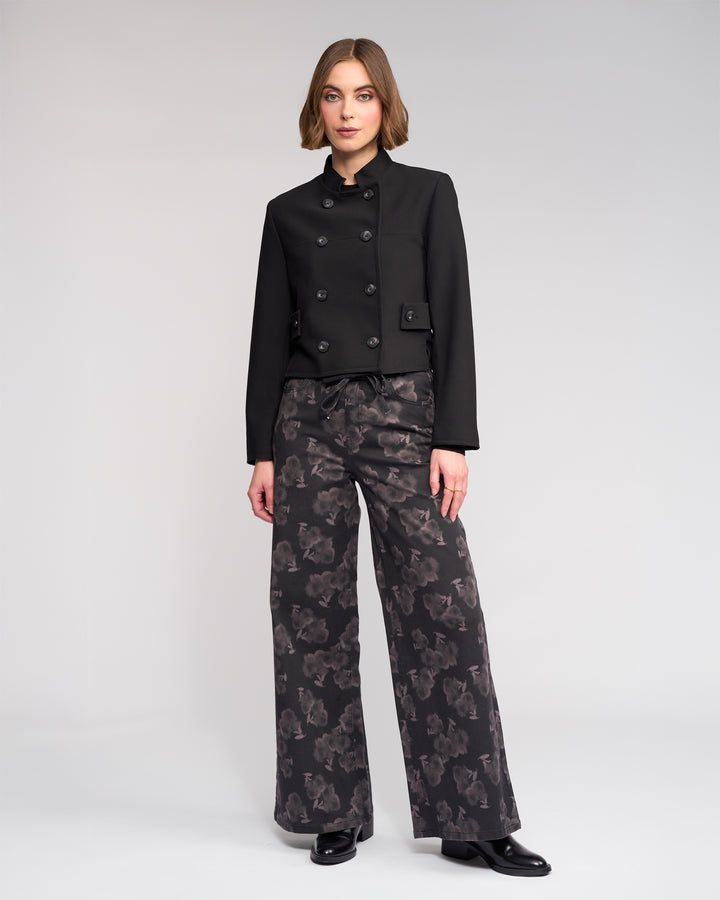 A woman with short, light brown hair and a neutral expression stands against a plain background wearing Knewe Label's Soho Jacket in black, paired with wide-leg patterned black trousers and black shoes.