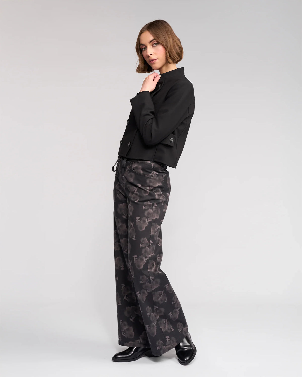 A woman with short brown hair models the Knewe Label Soho Jacket in black, posing in a studio with one hand near her chin against a plain light gray background. She pairs the jacket with wide-leg patterned pants and black shoes.