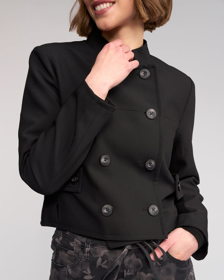 A woman smiles wearing the Soho Jacket - Black by Knewe Label, featuring large buttons, paired with black floral-patterned pants, against a light background.