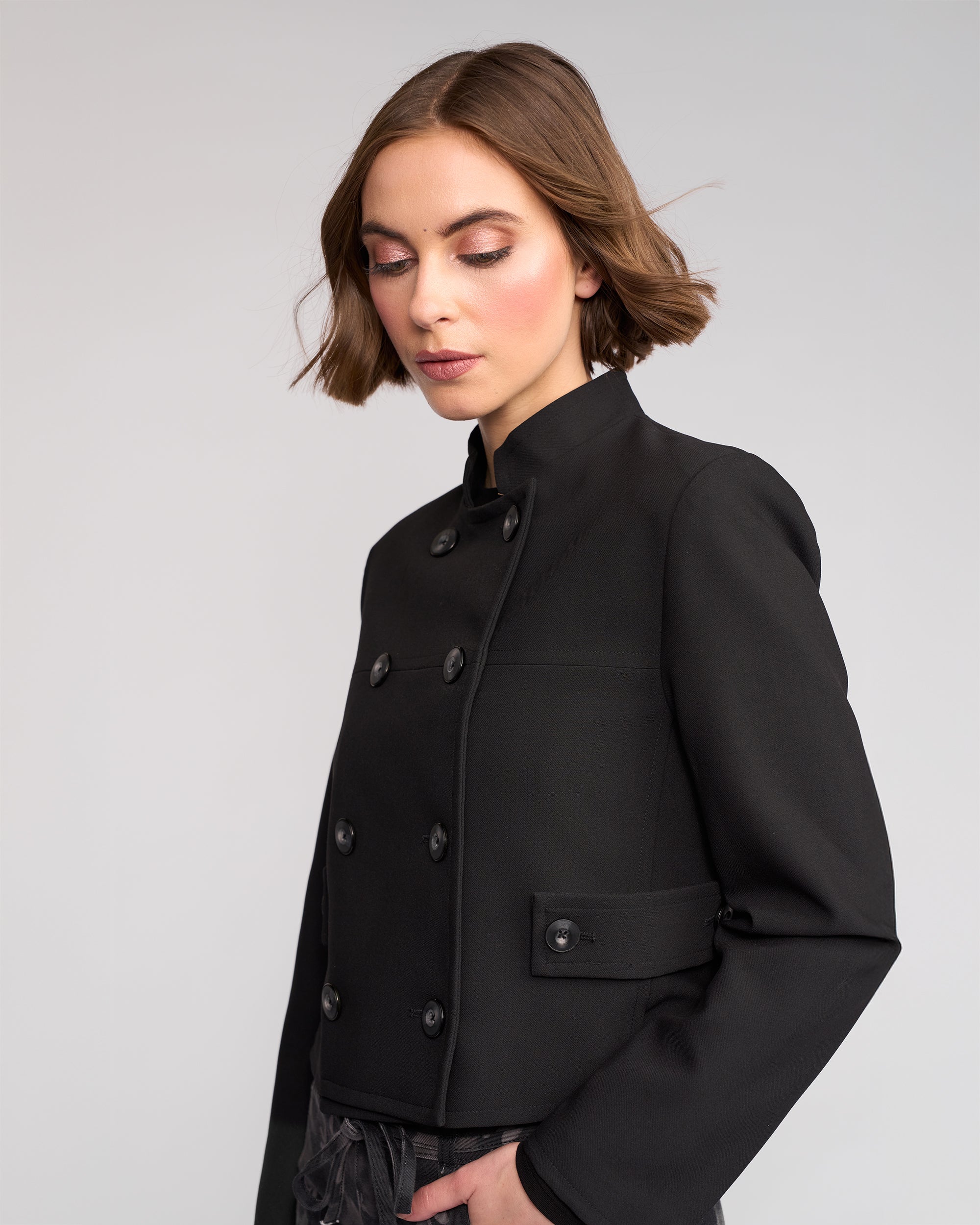 A woman with short brown hair wears the Soho Jacket - Black by Knewe Label. The fitted, double-breasted jacket features a high collar and lining. She stands against a plain light gray background, looking down with a calm expression.