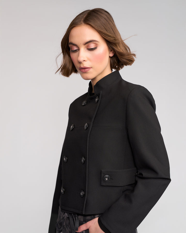 A woman with short brown hair wears the Soho Jacket - Black by Knewe Label. The fitted, double-breasted jacket features a high collar and lining. She stands against a plain light gray background, looking down with a calm expression.