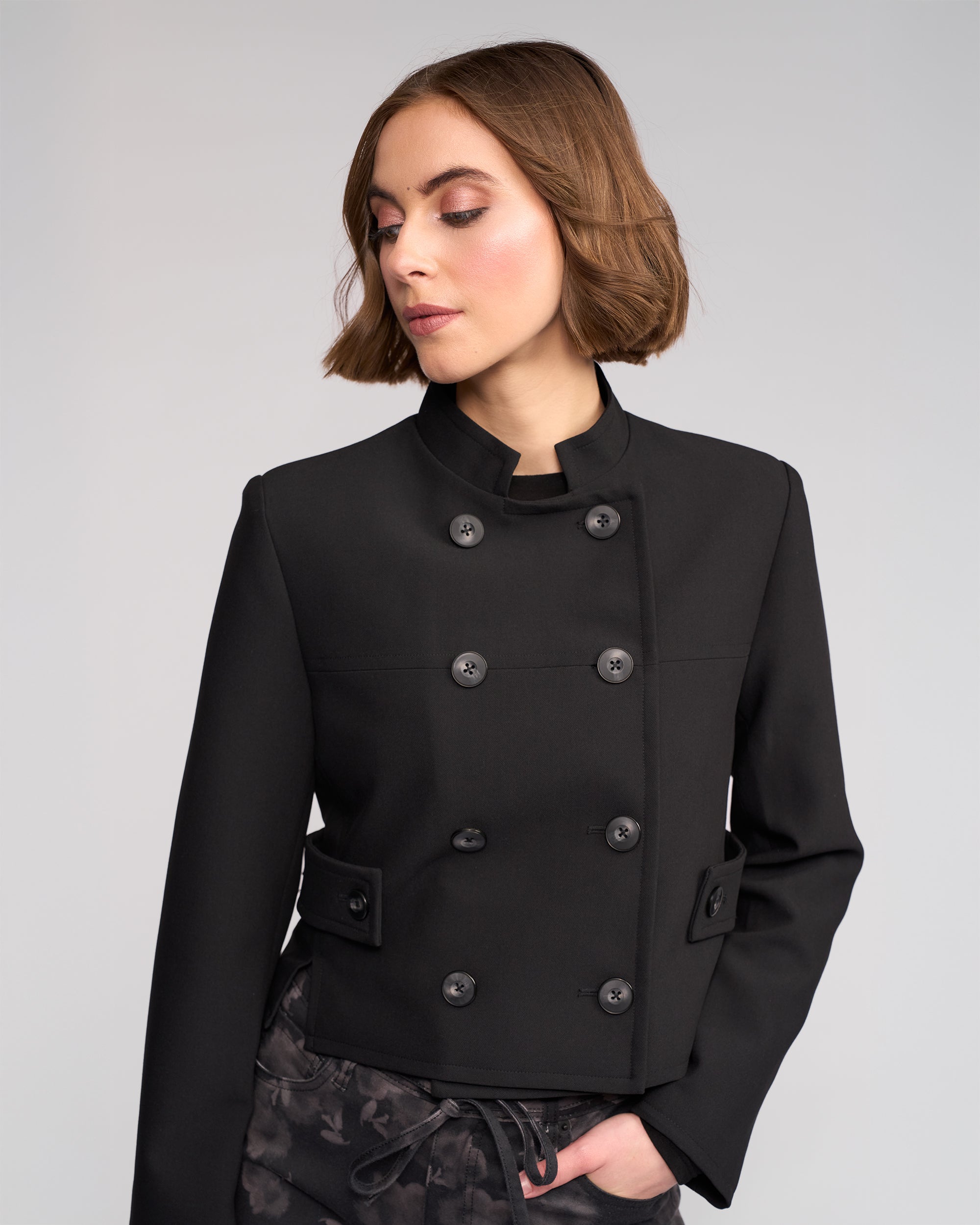 A woman with short brown hair wears the Knewe Label Soho Jacket in black, a double-breasted, cropped style with button details, looking down and slightly to the side against a plain light gray background.