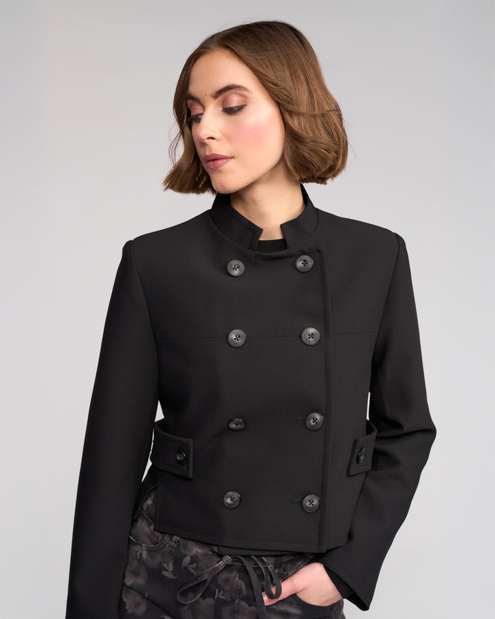 A woman with short brown hair wears the Knewe Label Soho Jacket in black, a double-breasted, cropped style with button details, looking down and slightly to the side against a plain light gray background.