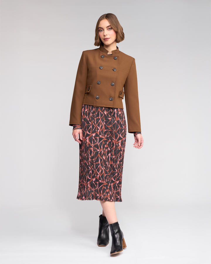 A woman with short, wavy brown hair confidently poses against a plain light gray background, wearing the Knewe Label Soho Jacket in Cedar over a patterned midi dress and black ankle boots.